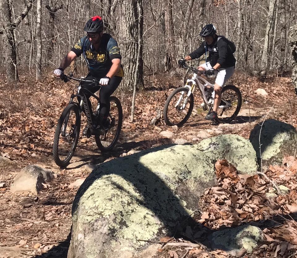 Two mountain bikers navigate a rocky trail through a wooded area in early spring. One rider is dressed in dark attire and a helmet, while the other wears a black jacket and white shorts. The ground is covered with fallen leaves, and large rocks are scattered along the path. Trees without leaves form a backdrop against a clear sky. Coldwater Mountain mountain bike trail.