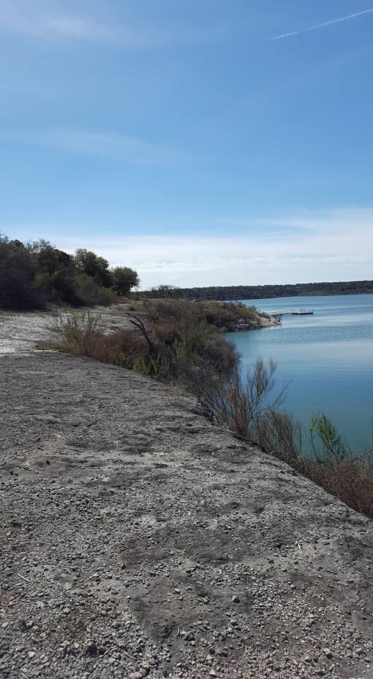 A tranquil lakeside scene with a gravel path leading to water, surrounded by sparse vegetation and distant trees under a clear blue sky. Goodwater Trail mountain bike trail.