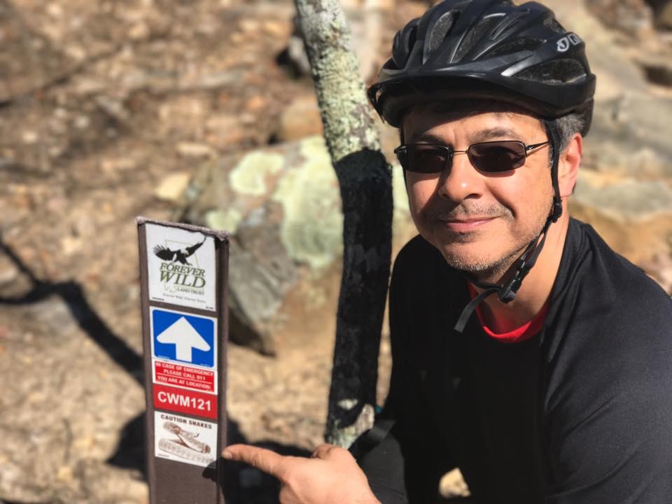 A person wearing a bicycle helmet and sunglasses smiles while pointing at a trail marker that features a "Forever Wild" sign, directional arrow, and caution about snakes. The background shows a natural, rocky landscape indicative of a hiking or biking trail. Coldwater Mountain mountain bike trail.