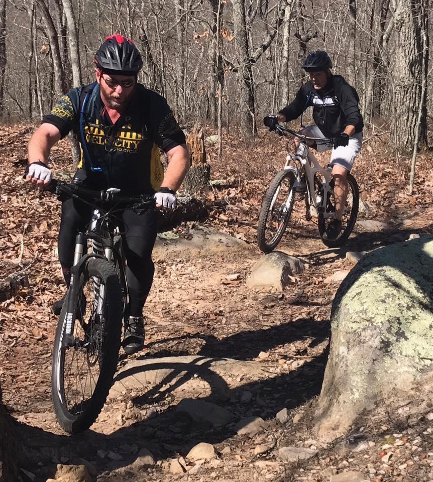 Two mountain bikers navigating a rocky trail in a wooded area. One cyclist is in the foreground, wearing a black jersey and helmet, concentrating as he maneuvers over a boulder. The second cyclist is in the background, wearing a black and white jacket and shorts, riding on a path covered with fallen leaves and stones. The trees in the background are bare, typical of a winter landscape. Coldwater Mountain mountain bike trail.
