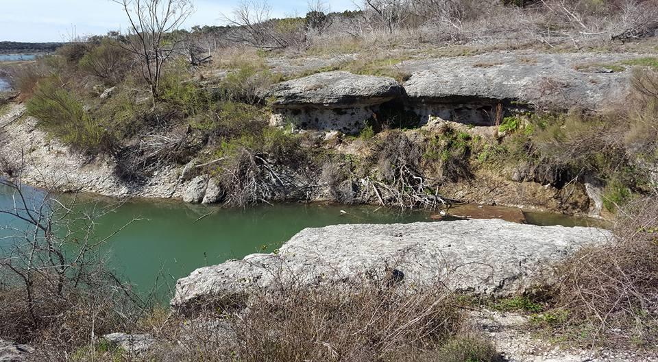 A rocky shoreline with green water reflecting the landscape. The area features large boulders and patches of vegetation, alongside dry branches and shrubs, set against a backdrop of sparse trees and a clear blue sky. Goodwater Trail mountain bike trail.