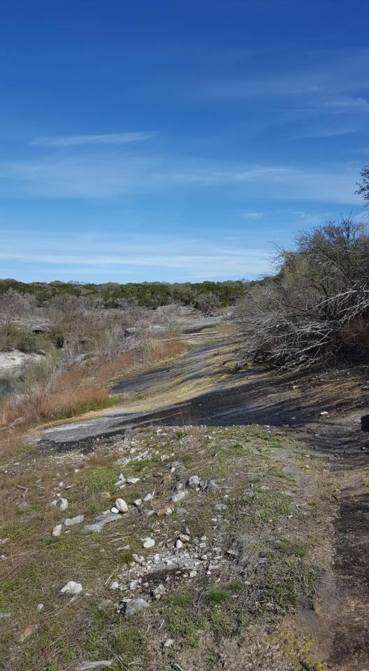 A scenic view of a dry, rocky landscape under a clear blue sky, featuring sparse vegetation, including grasses and bare trees. A winding path can be seen leading through the area, indicating possible human activity in the natural setting. Goodwater Trail mountain bike trail.