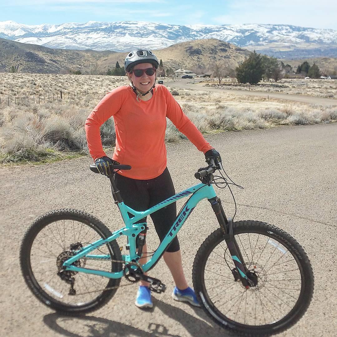Trek Remedy 8 27.5/650b: A person in athletic wear smiles while standing beside a turquoise mountain bike on a gravel road. They are wearing a black helmet and sunglasses, with a backdrop of snow-capped mountains and a clear blue sky. The surrounding landscape features dry grass and sparse vegetation.