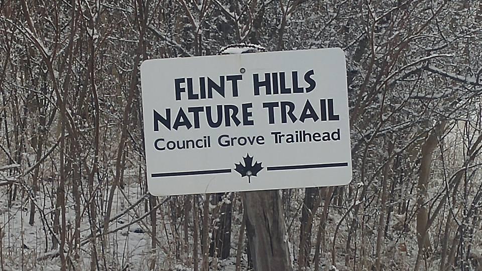 Sign for the Flint Hills Nature Trail at the Council Grove Trailhead, with a snowy background and surrounded by trees. Flint Hills Nature Trail (Rail Trail) mountain bike trail.