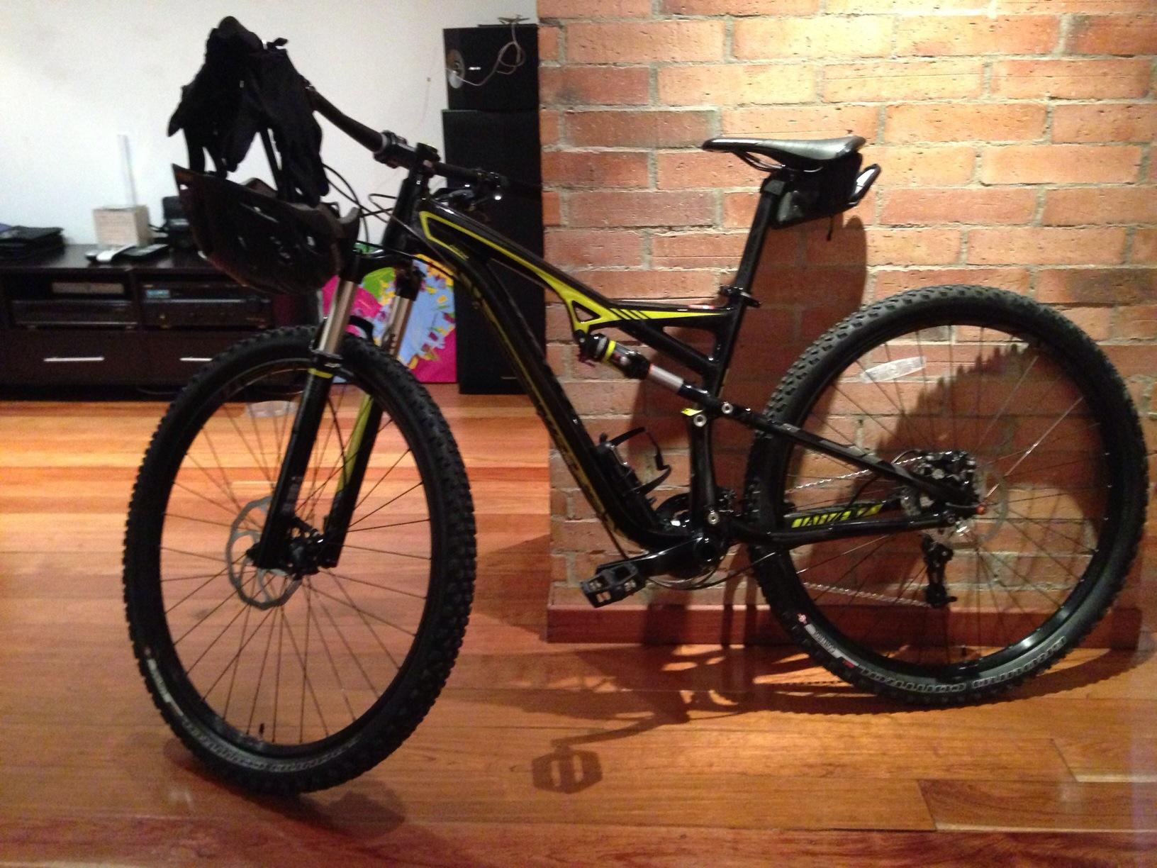Specialized Camber Comp 29: A black and yellow mountain bike leaning against a brick wall, with a decorative basket attached to the front and gloves draped over the handlebars. The floor is wooden, and a dark cabinet is visible in the background.