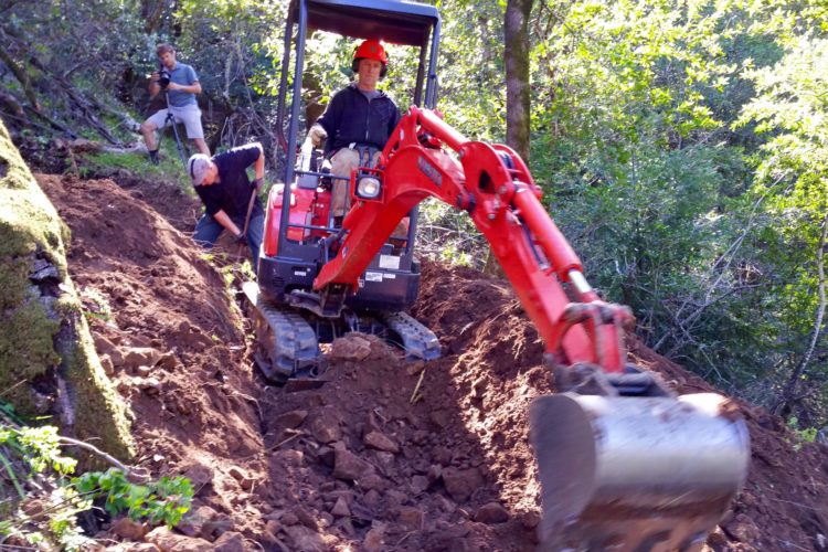 A small excavator is digging into the earth on a sloped terrain in a forested area. A person is operating the excavator while another individual is shoveling dirt nearby. A third person is capturing the scene with a camera. Surrounding greenery and sunlight filtering through the trees can be seen in the background.