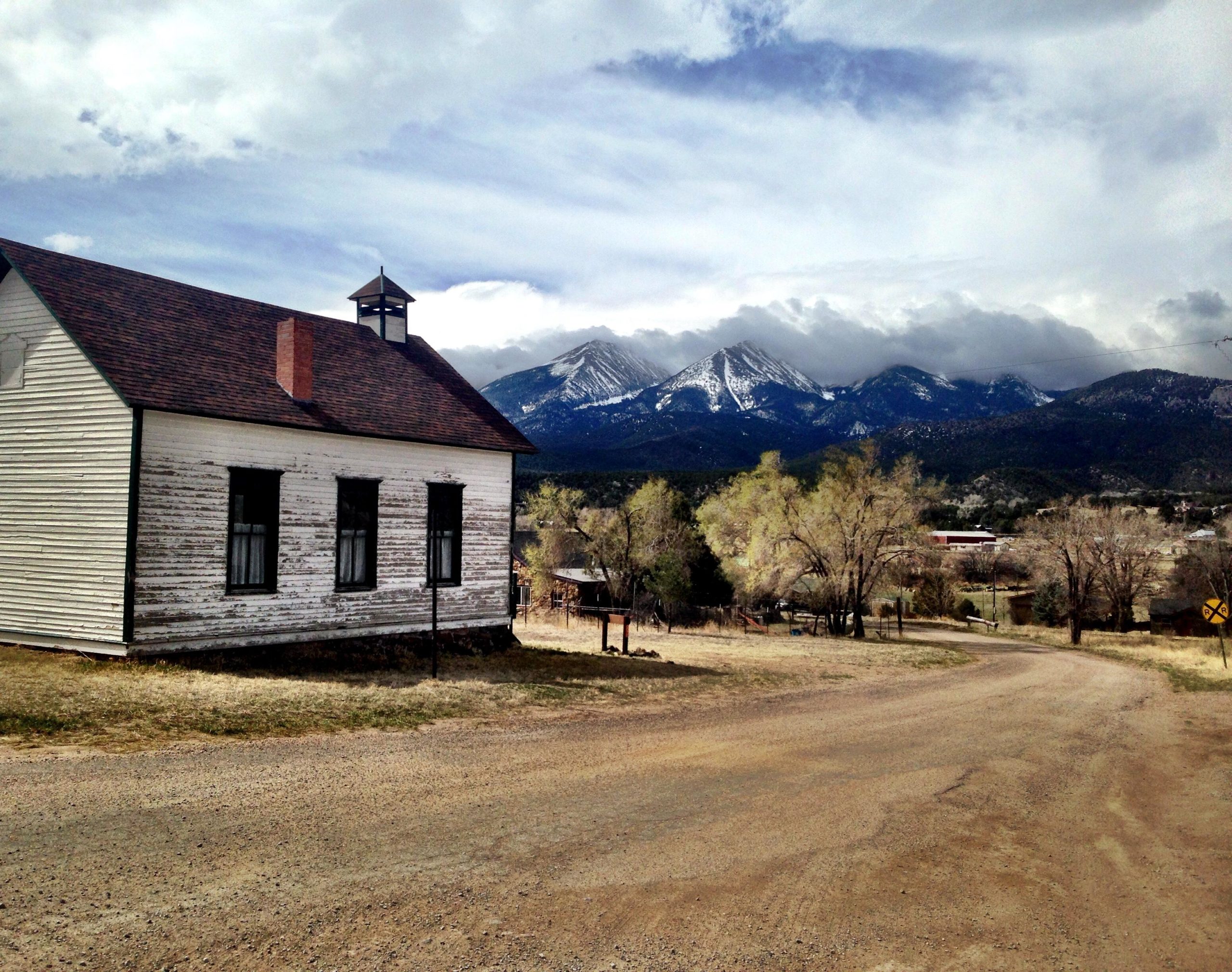 A rustic wooden building with a bell tower sits beside a dirt road, framed by snow-capped mountains in the background. The landscape features sparse trees and a cloudy sky, creating a serene rural atmosphere. Road #45 mountain bike trail.