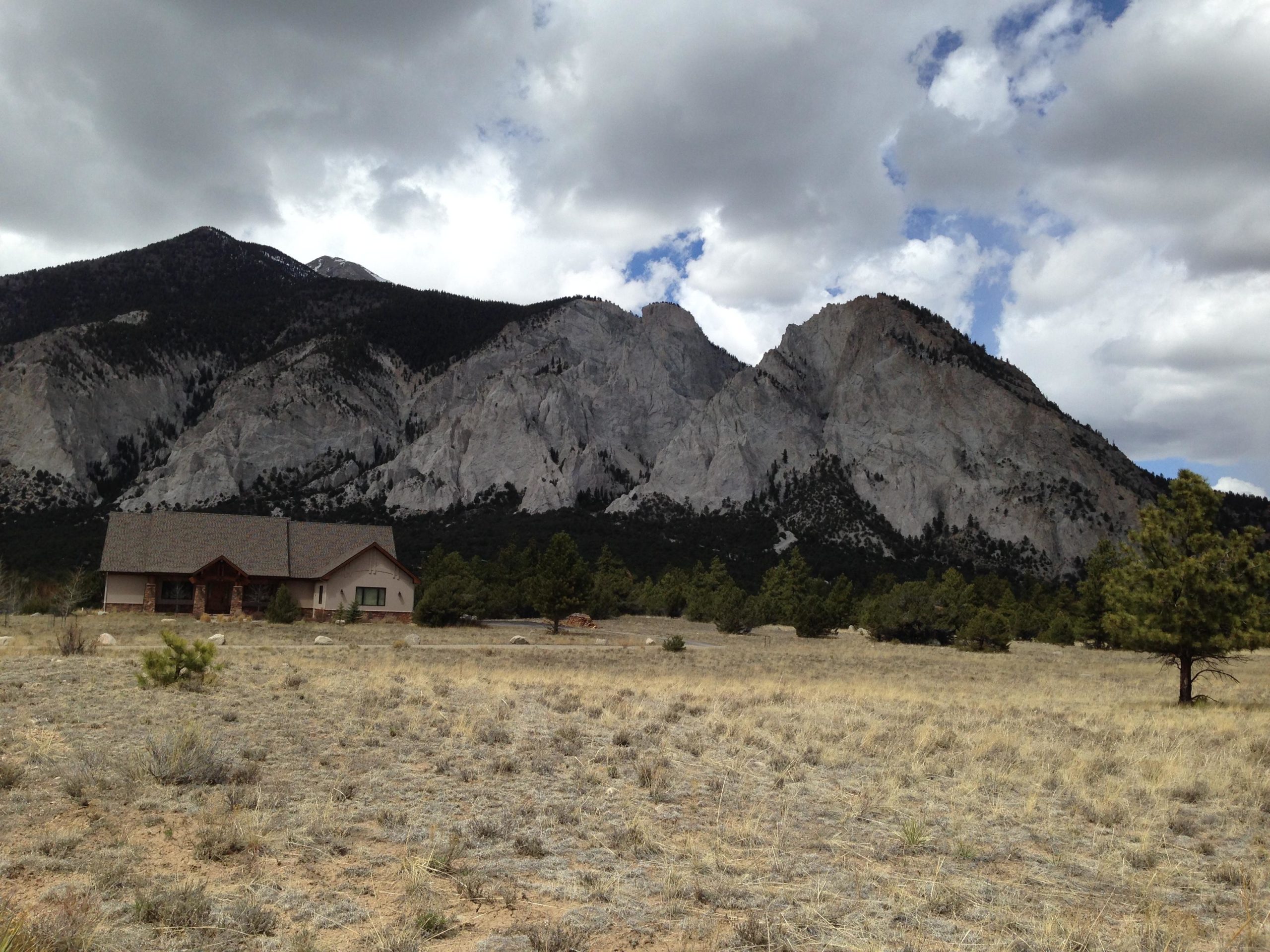 A picturesque landscape featuring a modern house in the foreground, surrounded by a grassy field and sparse trees. In the background, towering rocky mountains rise beneath a cloudy sky, creating a dramatic natural setting. Road #289 mountain bike trail.