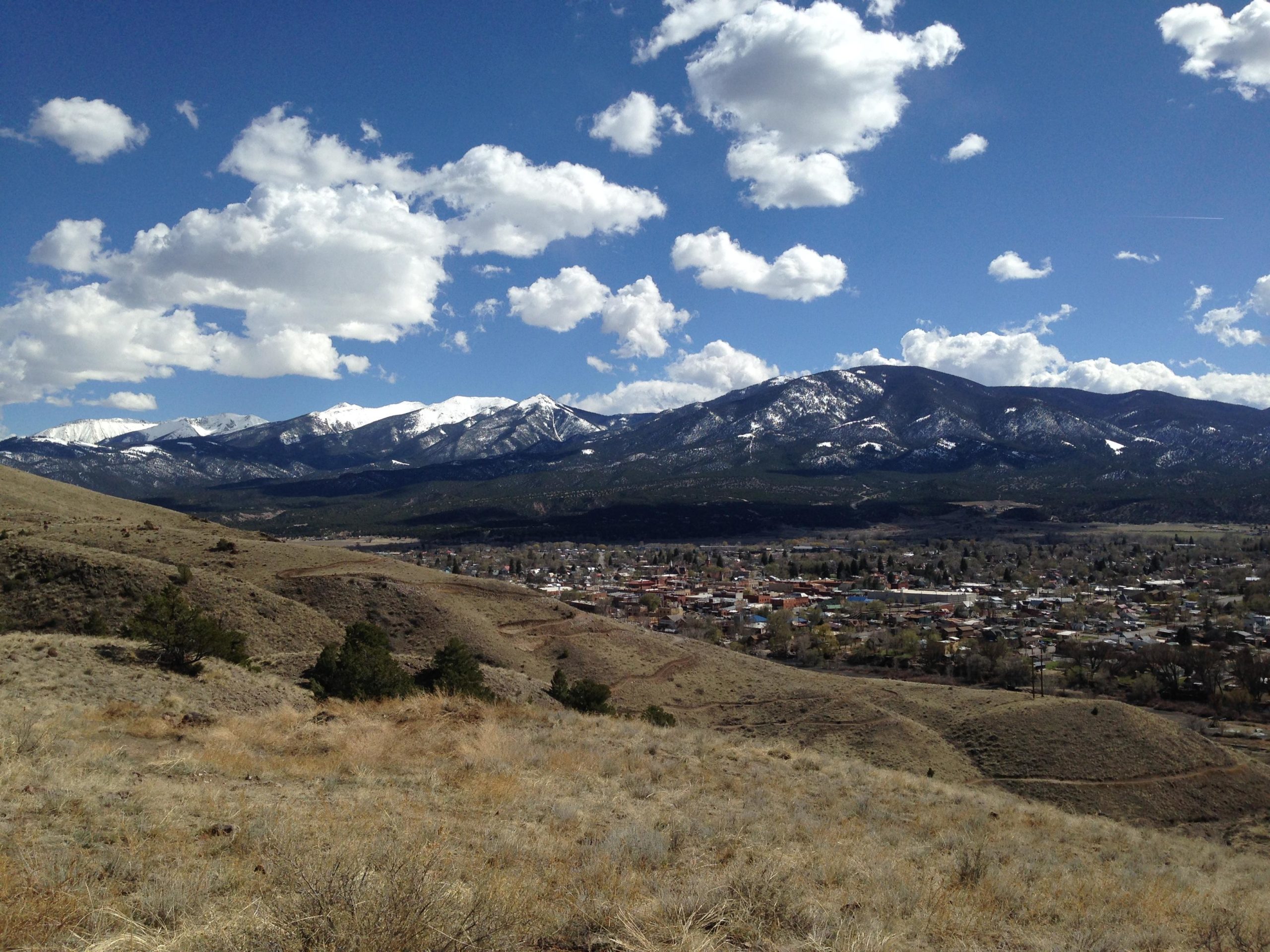 A panoramic view of a mountainous landscape featuring snow-capped peaks under a blue sky dotted with fluffy white clouds. In the foreground, dry grass and sparse vegetation cover the rolling hills, while a small town is nestled in the valley below, surrounded by trees and hills. Arkansas Hills mountain bike trail.