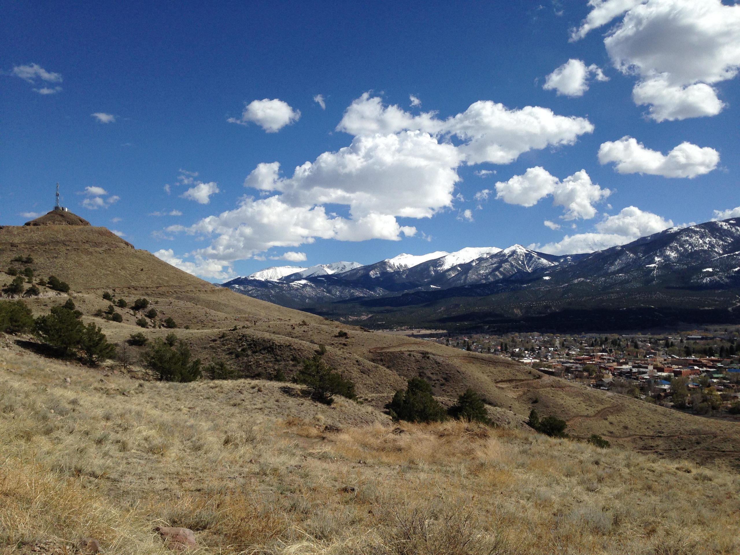 A scenic view of rolling hills and snow-capped mountains under a bright blue sky with fluffy white clouds. In the foreground, dry grass and small trees can be seen, while a small town is nestled in the valley below, showcasing a blend of nature and human settlement. Arkansas Hills mountain bike trail.