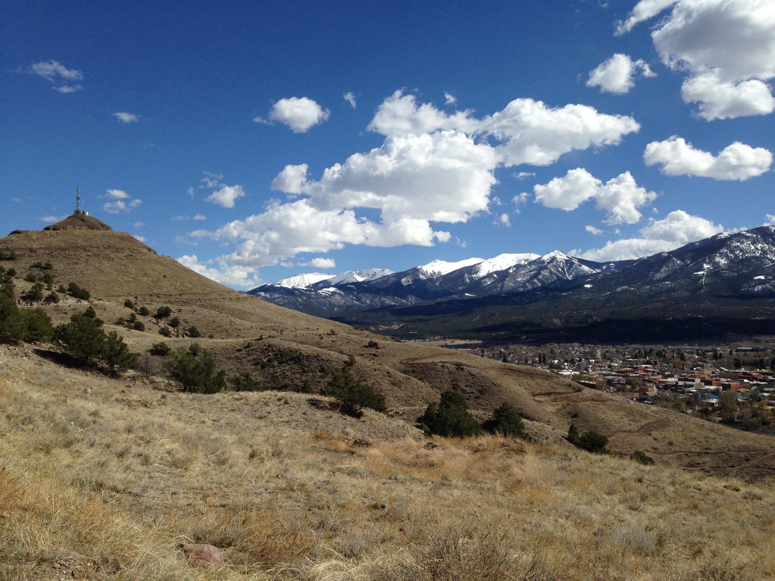 A scenic landscape featuring rolling hills and mountains under a blue sky with fluffy white clouds. In the foreground, there are grassy slopes and scattered pine trees. In the background, snow-capped mountains rise majestically, while a small town is visible in the valley below. A communications tower stands atop a nearby hill, contributing to the natural beauty of the area. Arkansas Hills mountain bike trail.