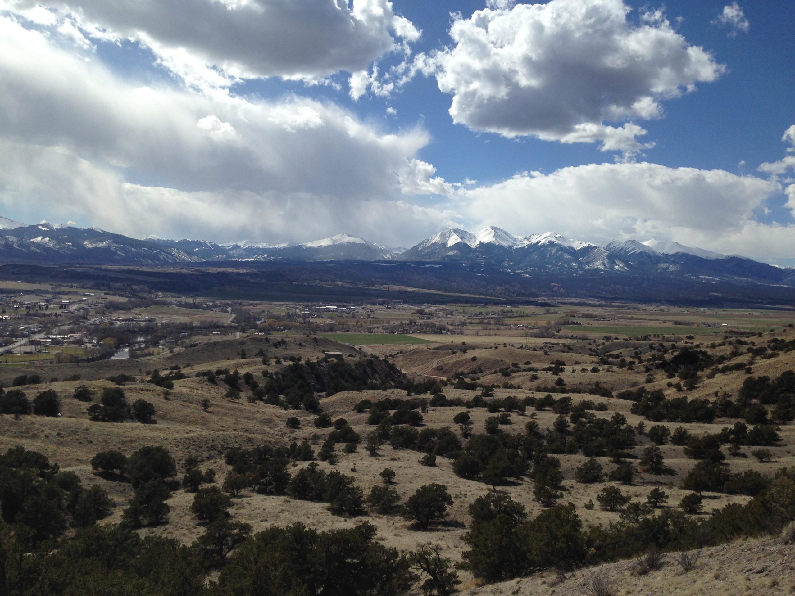 A panoramic view of a mountainous landscape under a partly cloudy sky. Snow-capped peaks rise in the background, while rolling hills and patches of greenery are visible in the foreground. A small river and a town can be seen in the valley below, showcasing the natural beauty of the region. Arkansas Hills mountain bike trail.