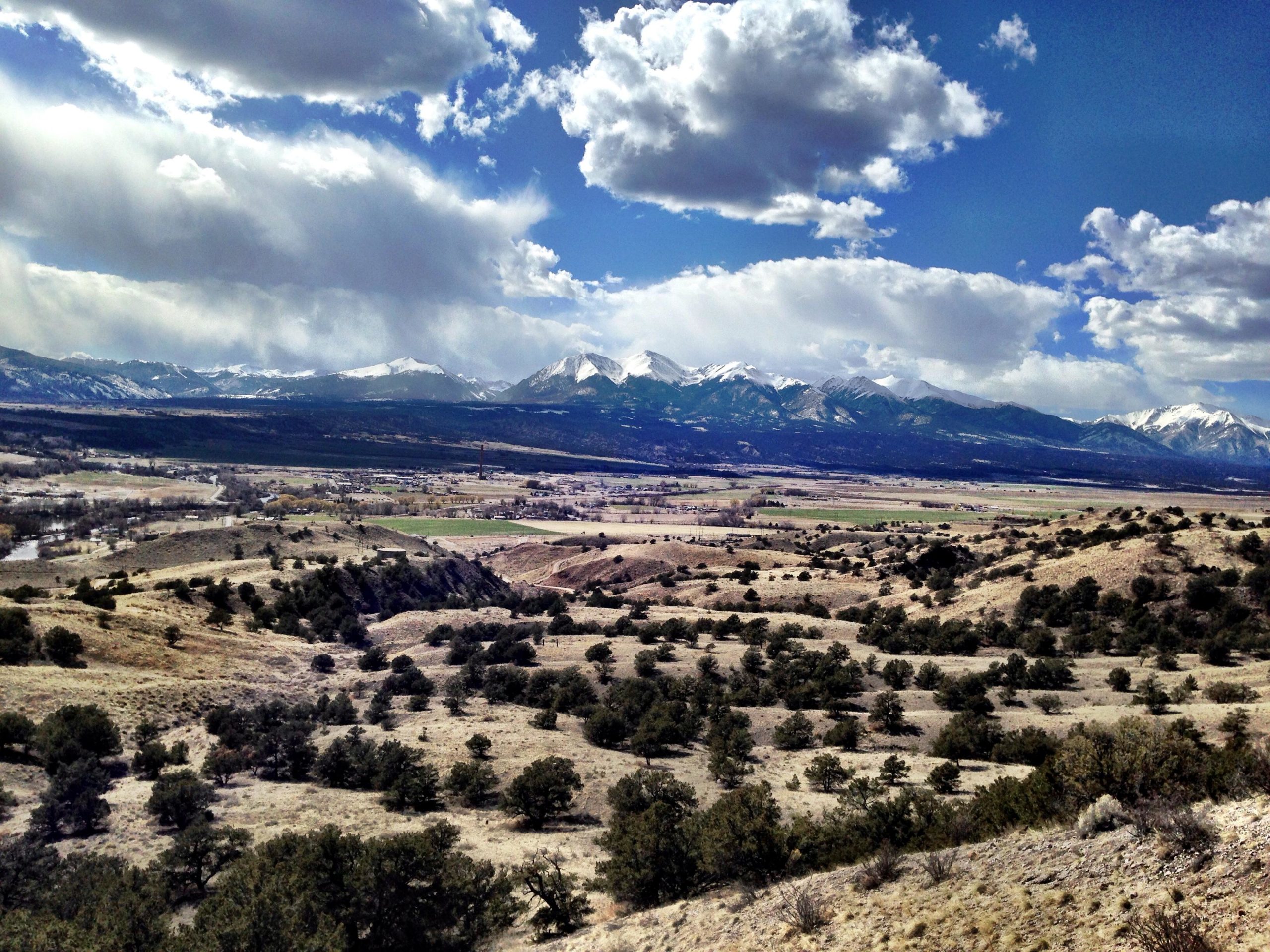 A panoramic view of a mountainous landscape featuring snow-capped peaks under a partly cloudy sky. In the foreground, rolling hills are dotted with small trees, while a broad valley extends into the distance, showcasing fields and winding rivers. The scene captures the natural beauty of the region, highlighting both rugged terrain and serene vistas. Arkansas Hills mountain bike trail.