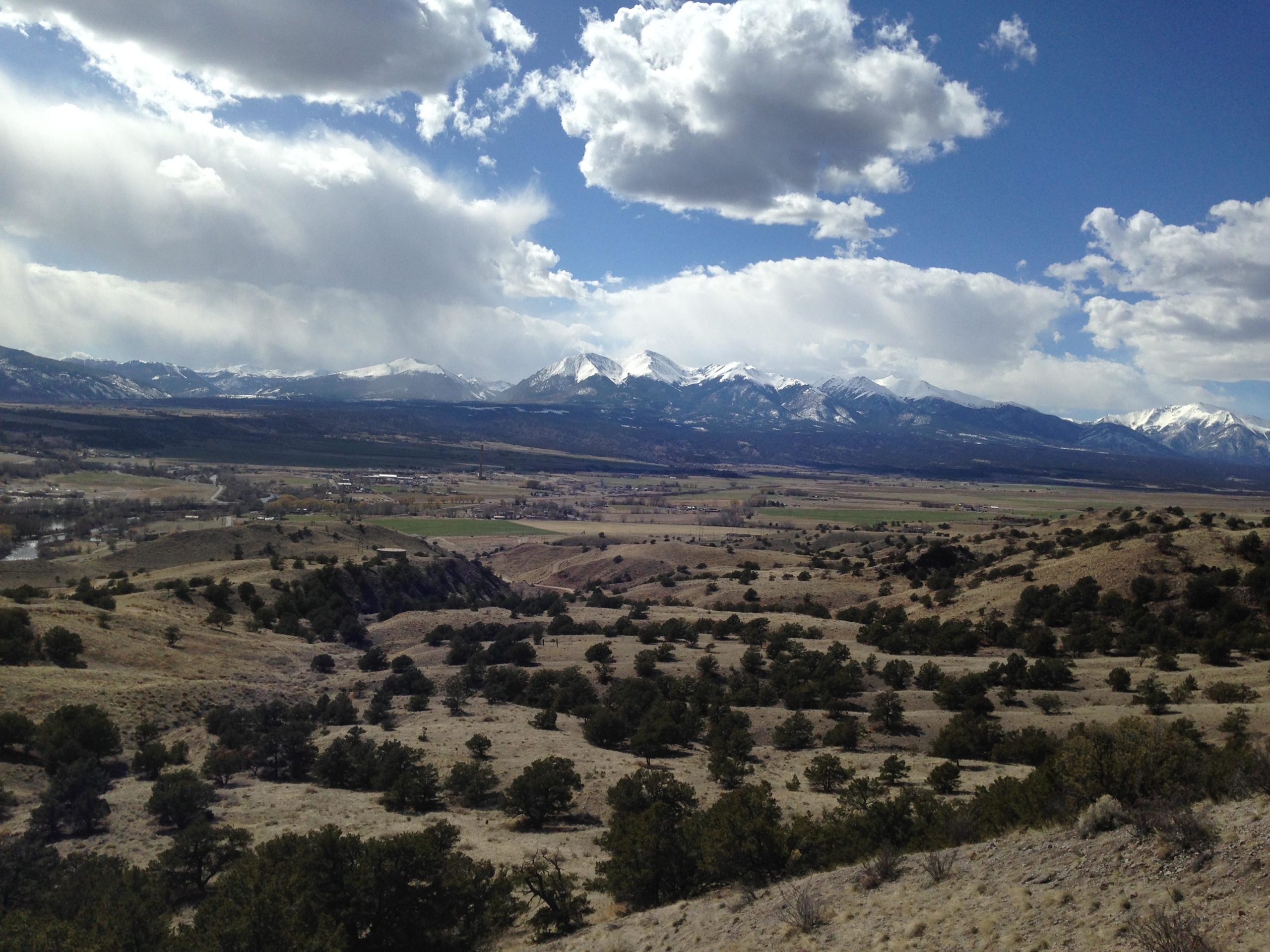 A panoramic view of a mountainous landscape under a partly cloudy sky, featuring snow-capped peaks in the background. In the foreground, rolling hills are dotted with evergreen trees and shrubs. A green valley stretches out below, showcasing patches of farmland and a winding river. The image captures the beauty of nature in a serene, open environment. Arkansas Hills mountain bike trail.