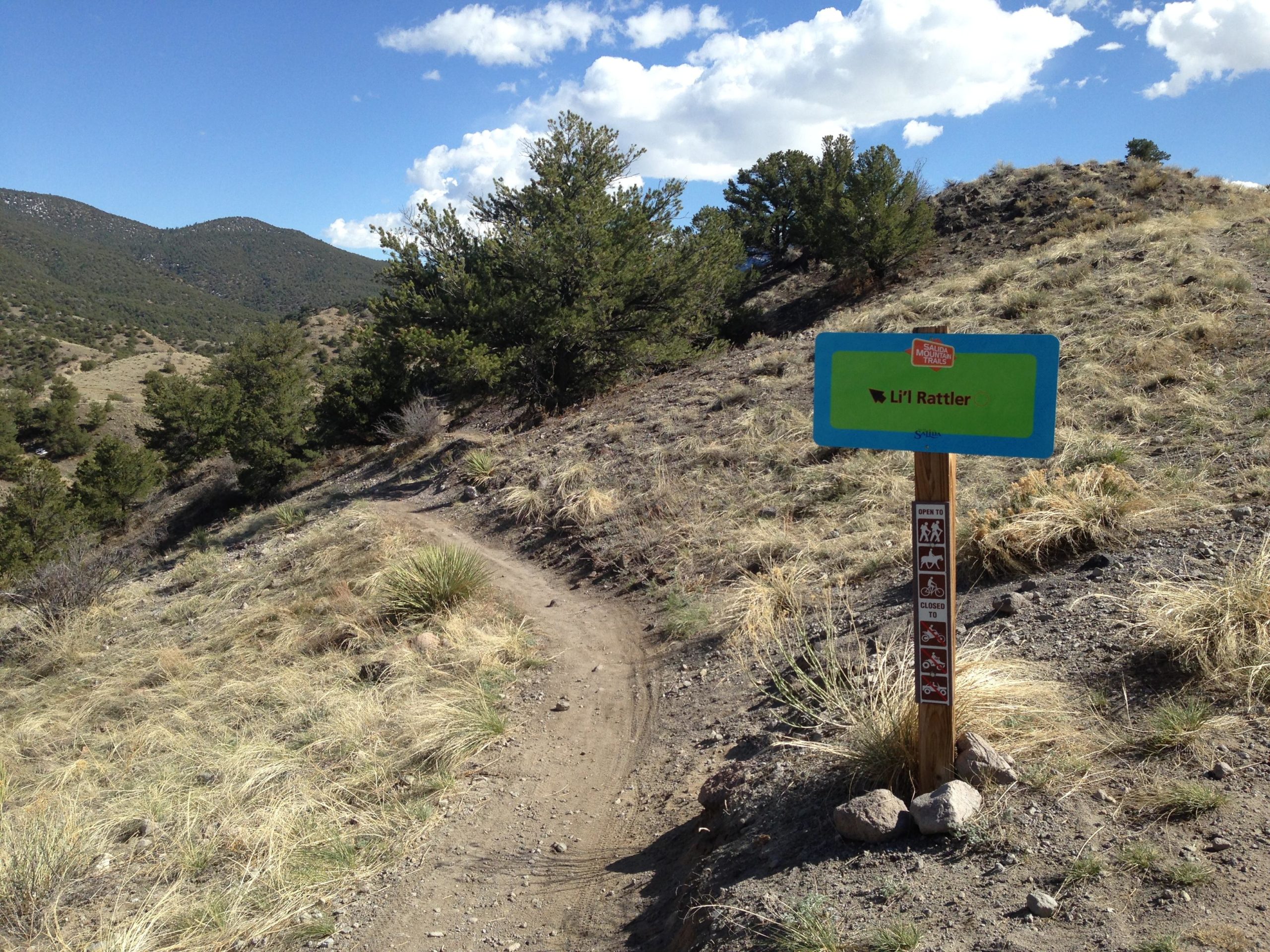 Sign marking the "Li'l Rattler" trail, surrounded by a dirt path and grassy terrain, with mountains in the background under a partly cloudy sky. Arkansas Hills mountain bike trail.