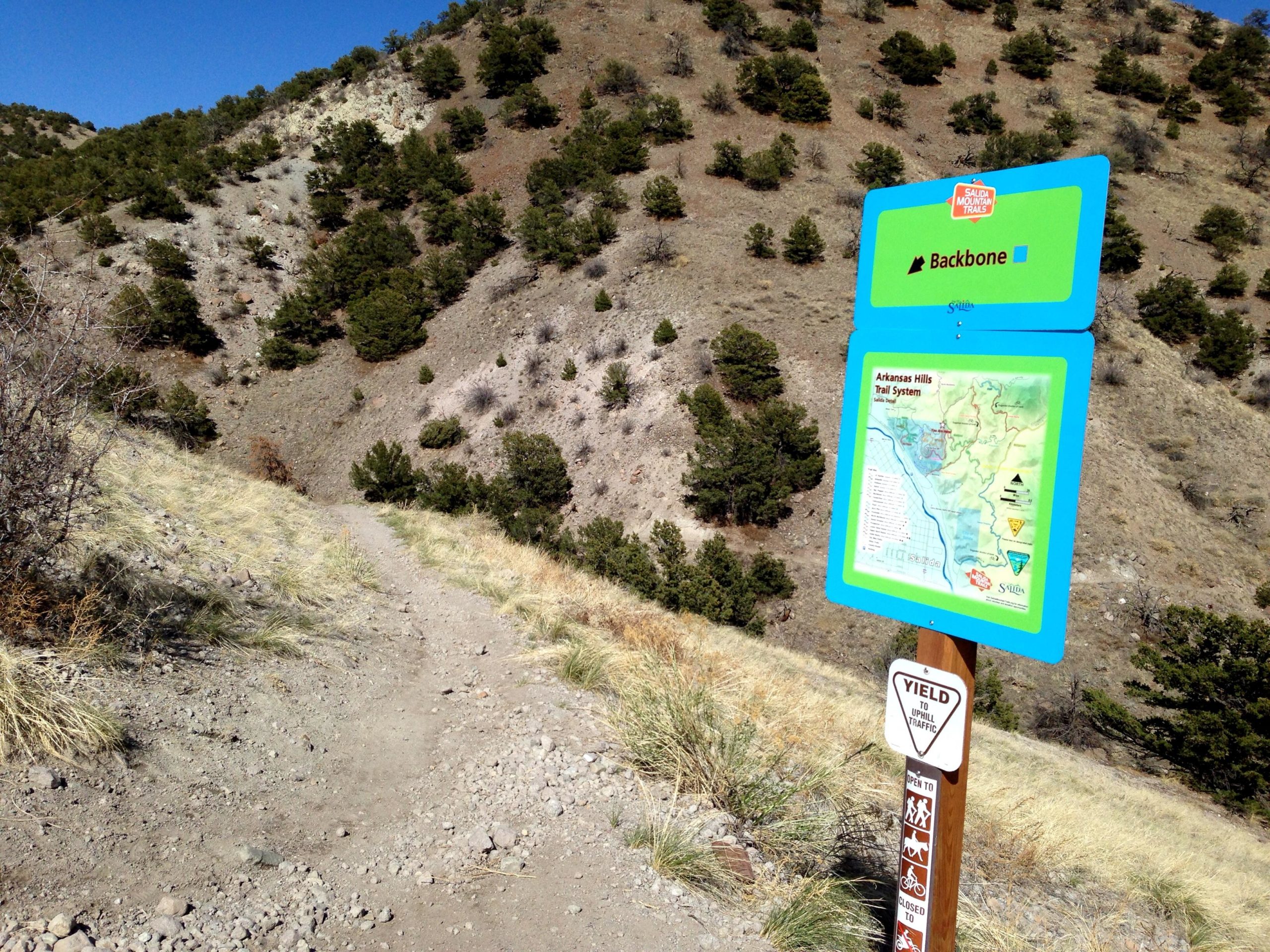 A scenic hiking trail with a dirt path leading upward, flanked by sparse vegetation and rocky terrain. In the foreground, a sign displays trail information, including the name "Backbone" and a map of the Arkansas Hills Trail System, along with a "Yield to Trail Traffic" warning for safety. The background features a slope covered with small trees and shrubs under a clear blue sky. Arkansas Hills mountain bike trail.