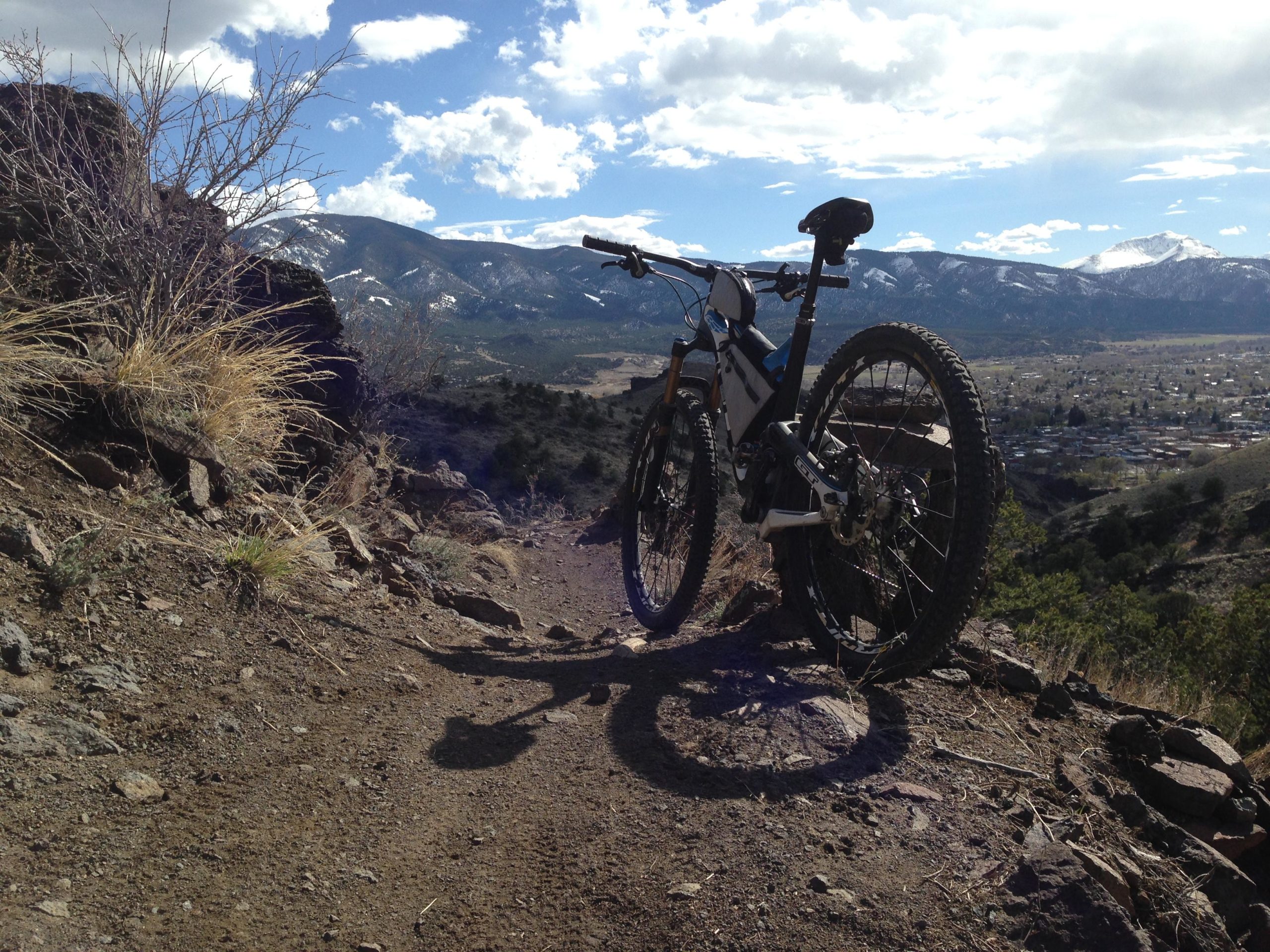 A mountain bike parked on a dirt trail overlooking a scenic landscape, with mountains in the background and a clear blue sky dotted with clouds. The foreground features rocky terrain and sparse vegetation, while a small town is visible in the valley below. Arkansas Hills mountain bike trail.