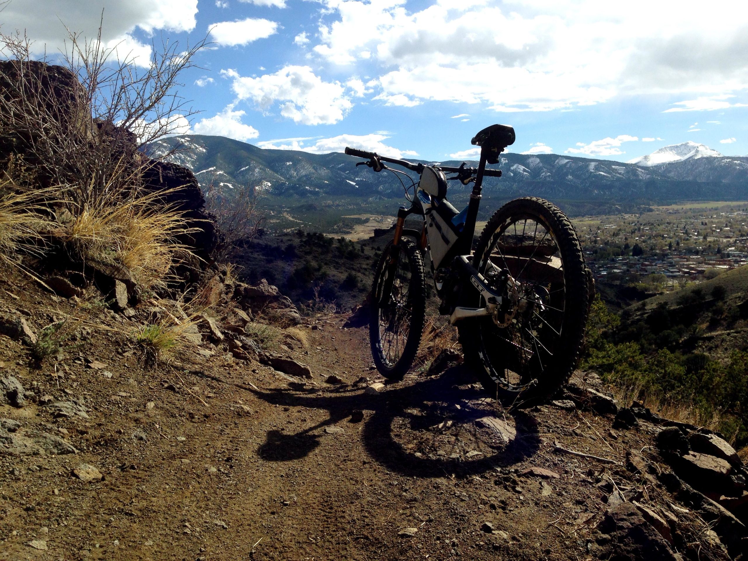 A mountain bike parked on a rocky trail overlooking a valley and distant snow-capped mountains under a partly cloudy sky. Arkansas Hills mountain bike trail.