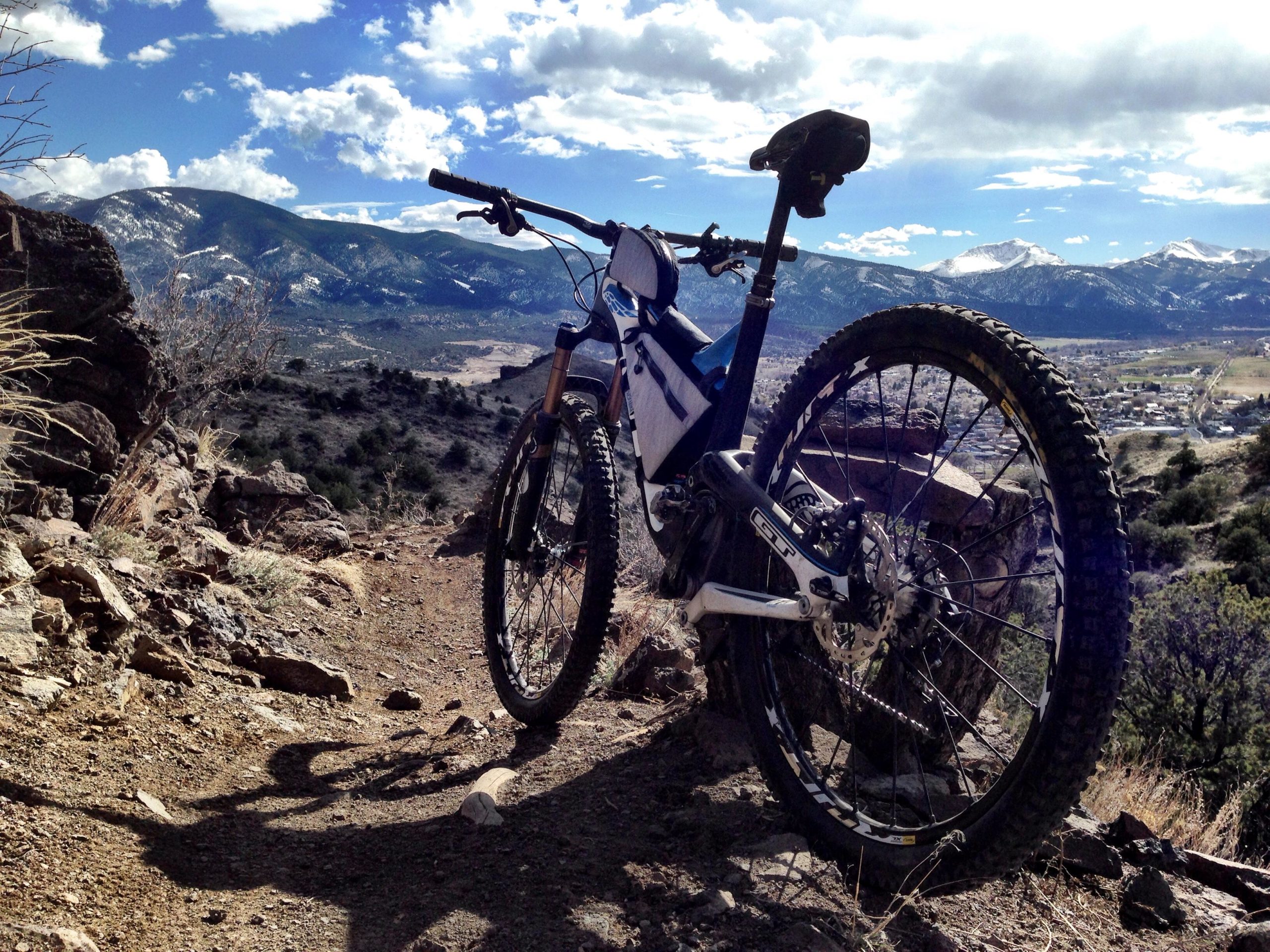 A mountain bike parked on a rocky trail overlooking a valley with mountains in the background and a partly cloudy sky. The bike is positioned to highlight its wheels and frame, with a scenic landscape stretching out behind it. Arkansas Hills mountain bike trail.