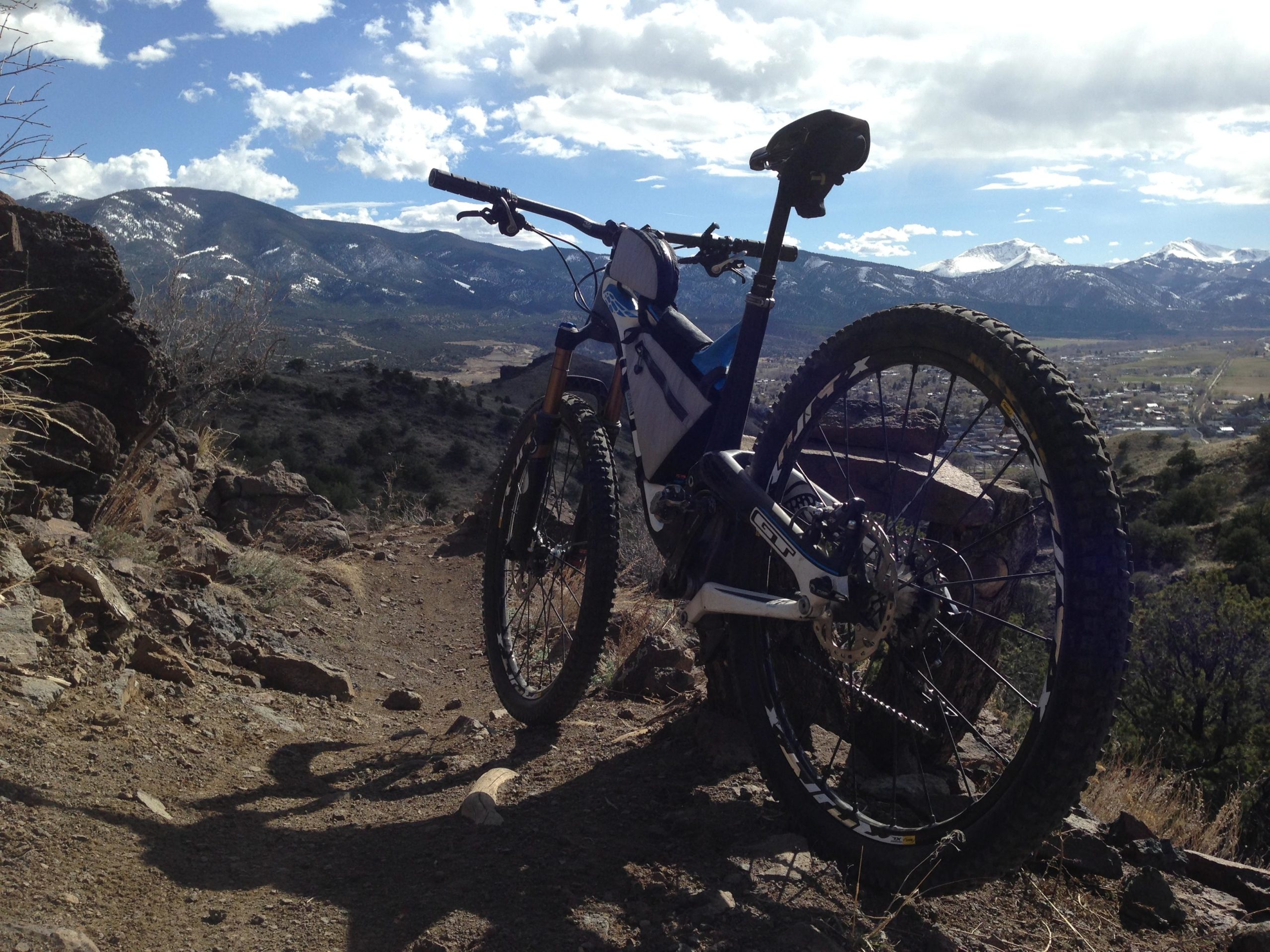 A mountain bike rests on a rocky trail with a scenic view of rolling hills and snow-capped mountains in the background. The sky is partly cloudy, showcasing a blend of blue and white hues. The foreground features dirt and grass, indicating an adventurous outdoor setting. Arkansas Hills mountain bike trail.