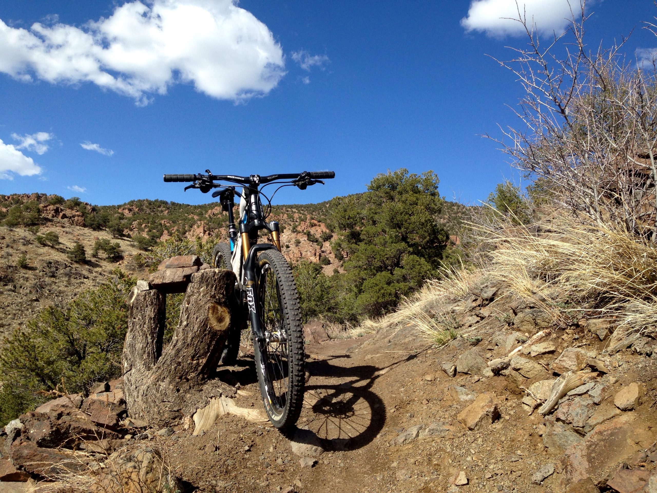 Mountain bike resting on a rocky trail, with a background of hills and blue sky dotted with clouds. A weathered tree stump is nearby, and sparse vegetation surrounds the area, highlighting an outdoor biking environment. Arkansas Hills mountain bike trail.