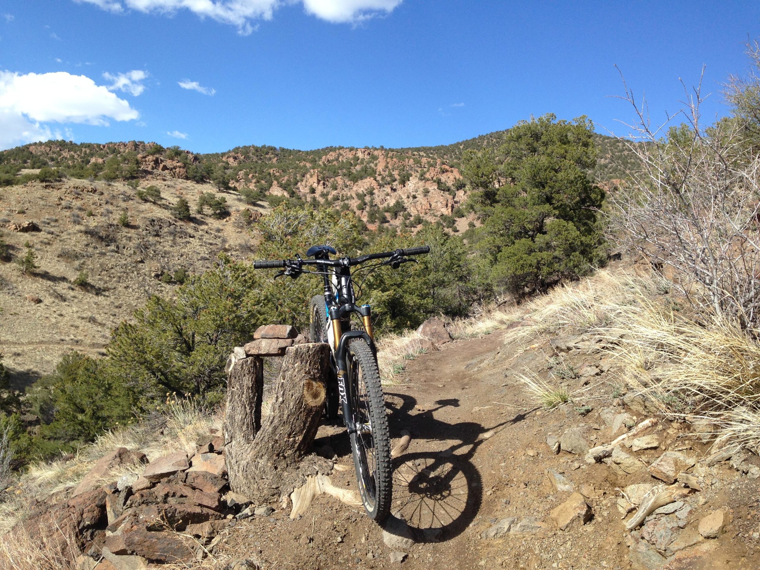 A mountain bike resting on a wooden stump along a rocky trail surrounded by hills and sparse vegetation, under a clear blue sky with a few clouds. Arkansas Hills mountain bike trail.