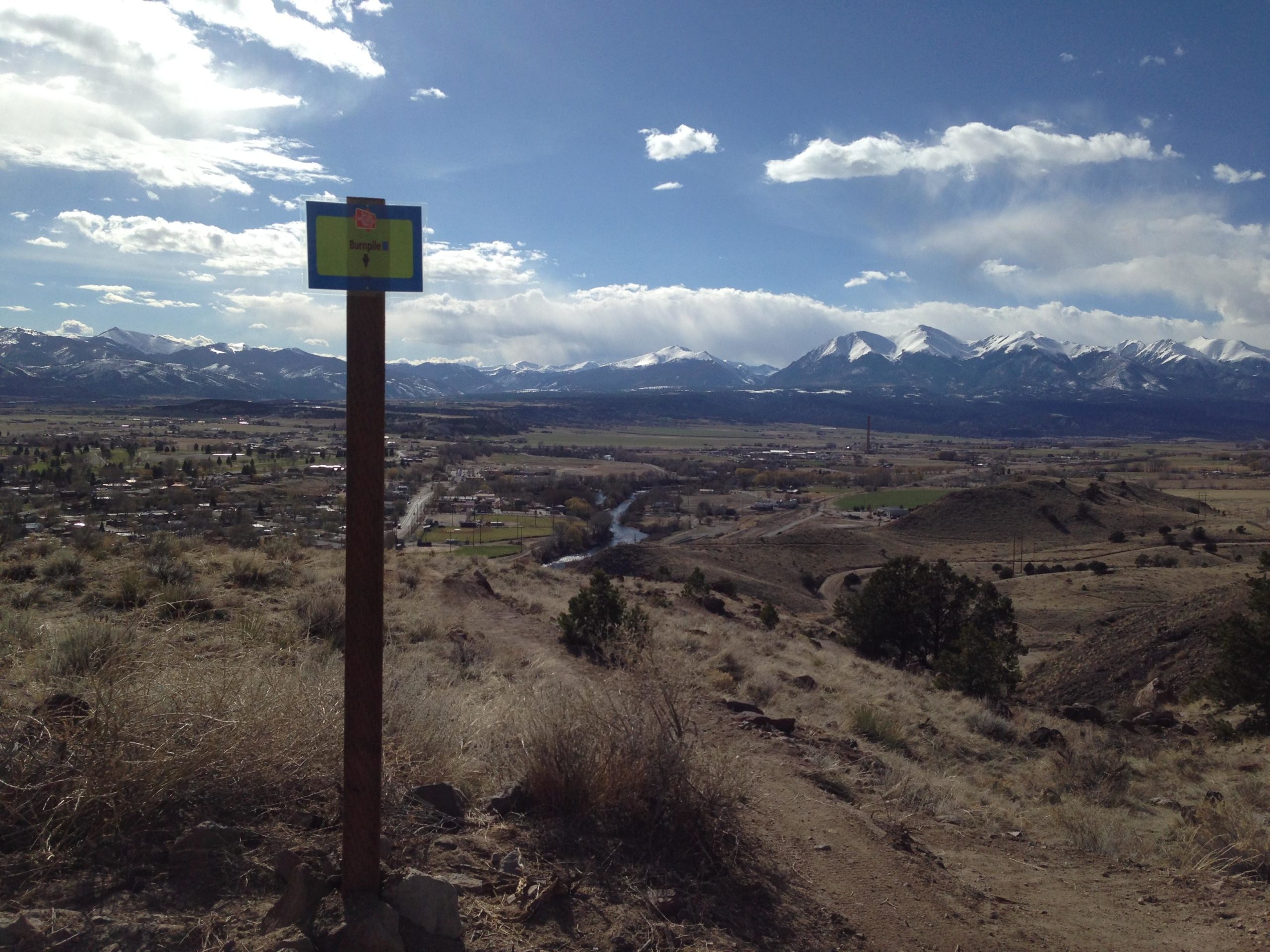 A scenic view of a valley with mountains in the background, featuring a signpost labeled “Bumpie III” in the foreground. The landscape includes rolling hills and a small town with winding roads and a river. The sky is partly cloudy, and the snow-capped peaks are visible in the distance, creating a picturesque outdoor setting. Arkansas Hills mountain bike trail.