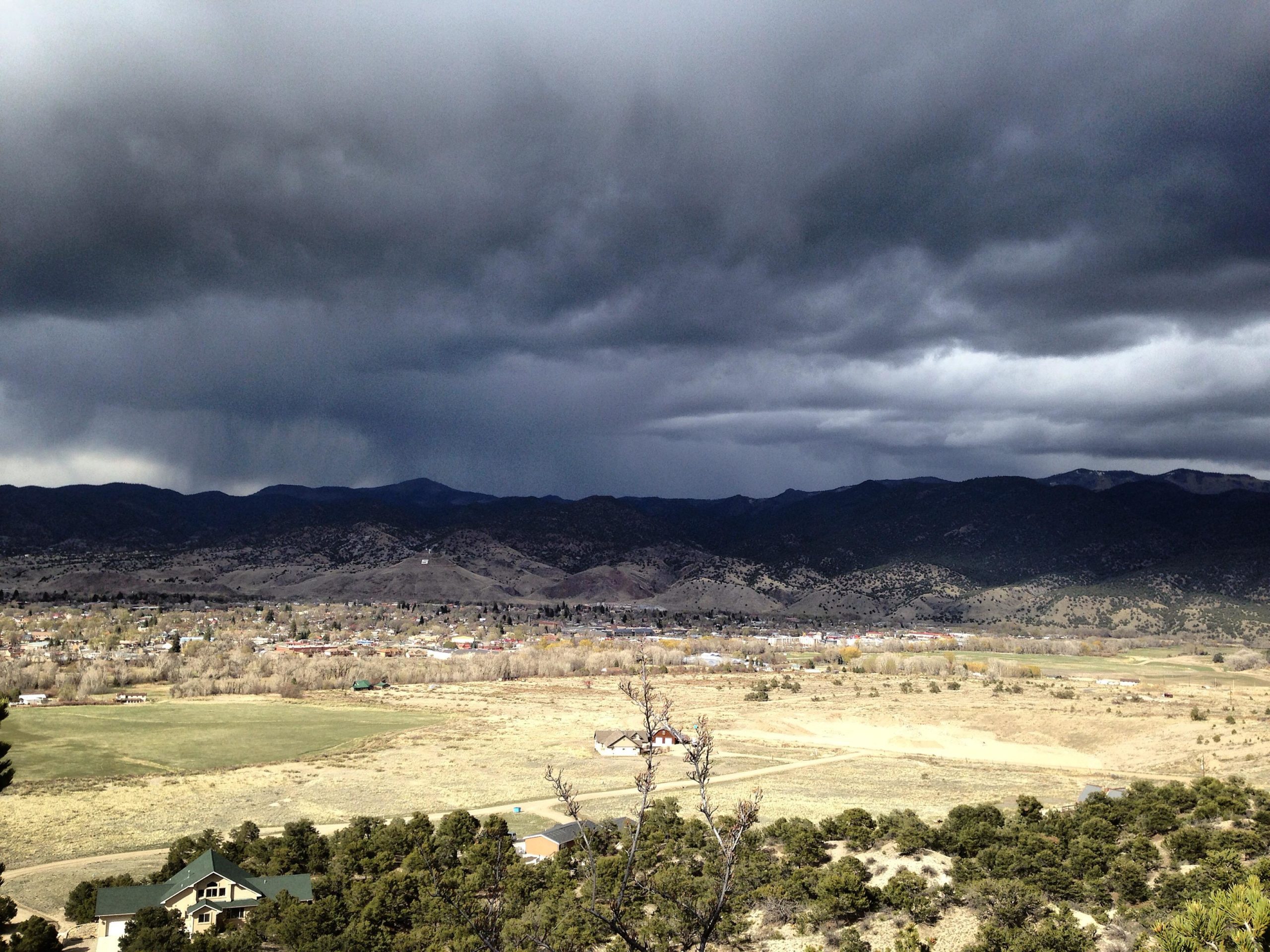 A panoramic view of a mountainous landscape under a dark, cloudy sky. The foreground features a patchwork of green fields and scattered homes, while the background showcases rolling hills and distant mountains. The atmosphere suggests an impending storm, with shades of gray dominating the sky. Methodist Mountain mountain bike trail.