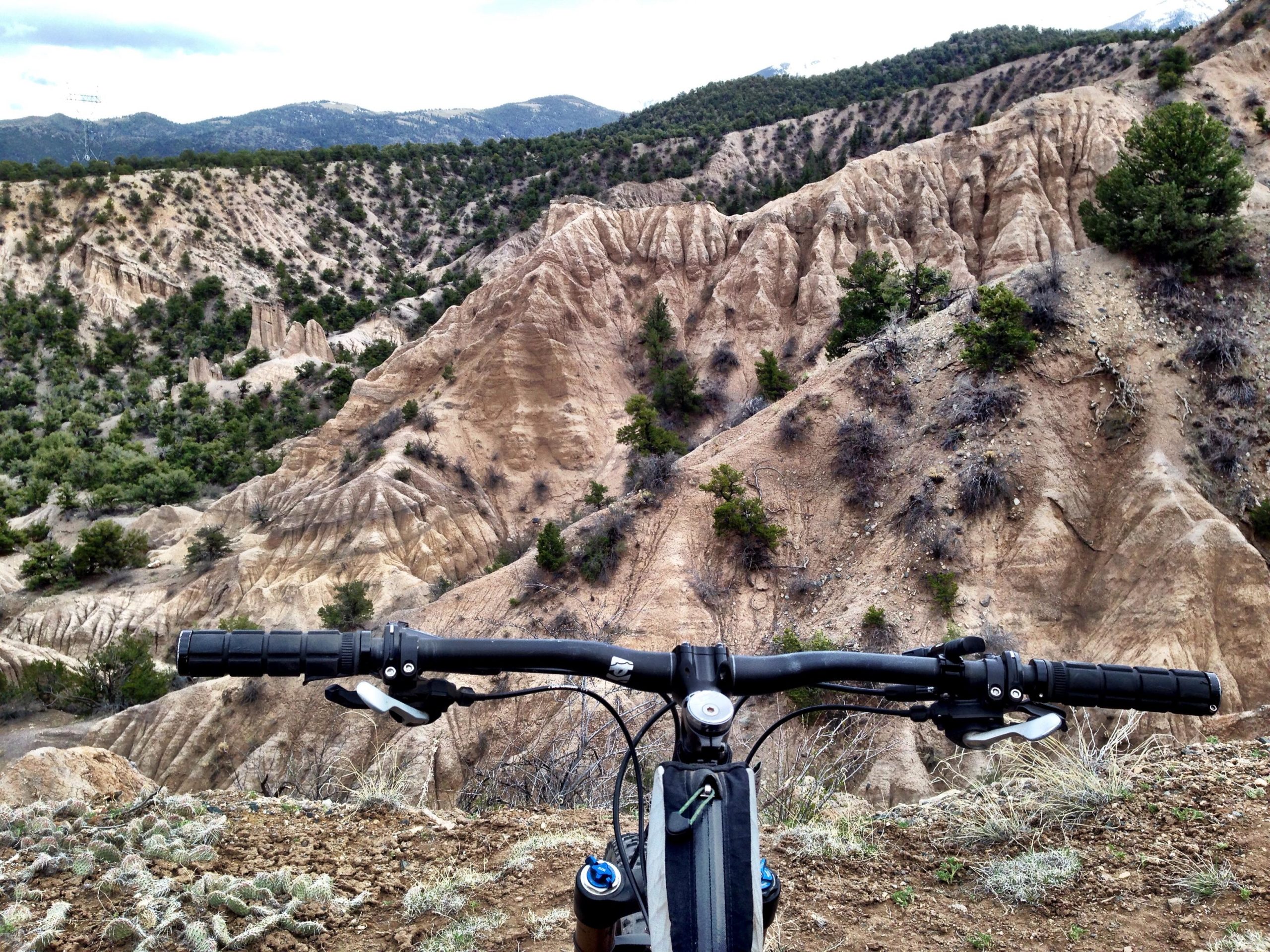 A mountain bike handlebar is shown in the foreground, overlooking a rugged, dry landscape with steep, sandy hills and sparse vegetation. The background features rolling mountains under a cloudy sky, creating a sense of adventure and exploration in nature. Methodist Mountain mountain bike trail.