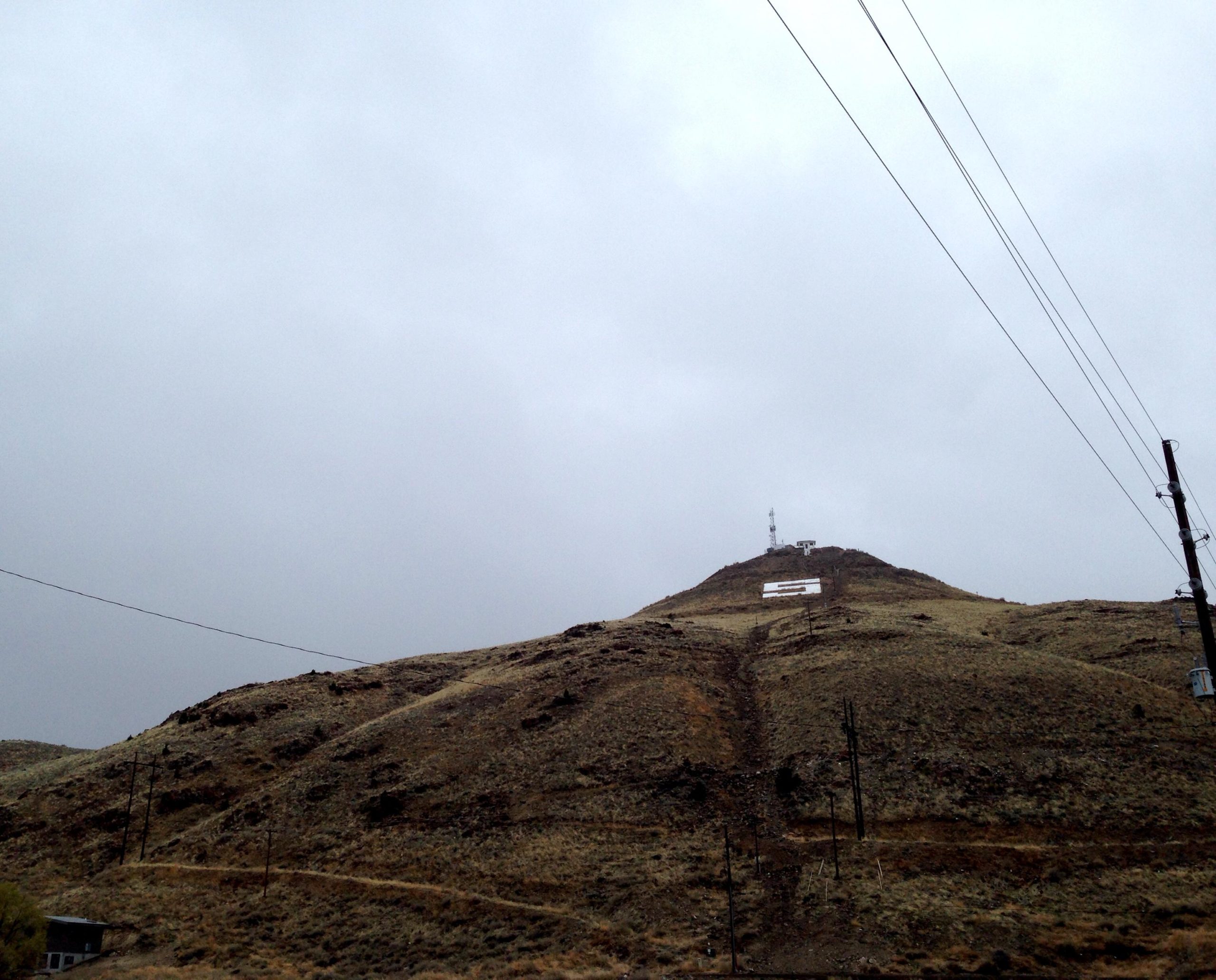 A hilly landscape under a cloudy sky, featuring a small building and a communication tower at the summit. Power lines are visible in the foreground, while the ground is mostly dry and rocky with sparse vegetation. Arkansas Hills mountain bike trail.