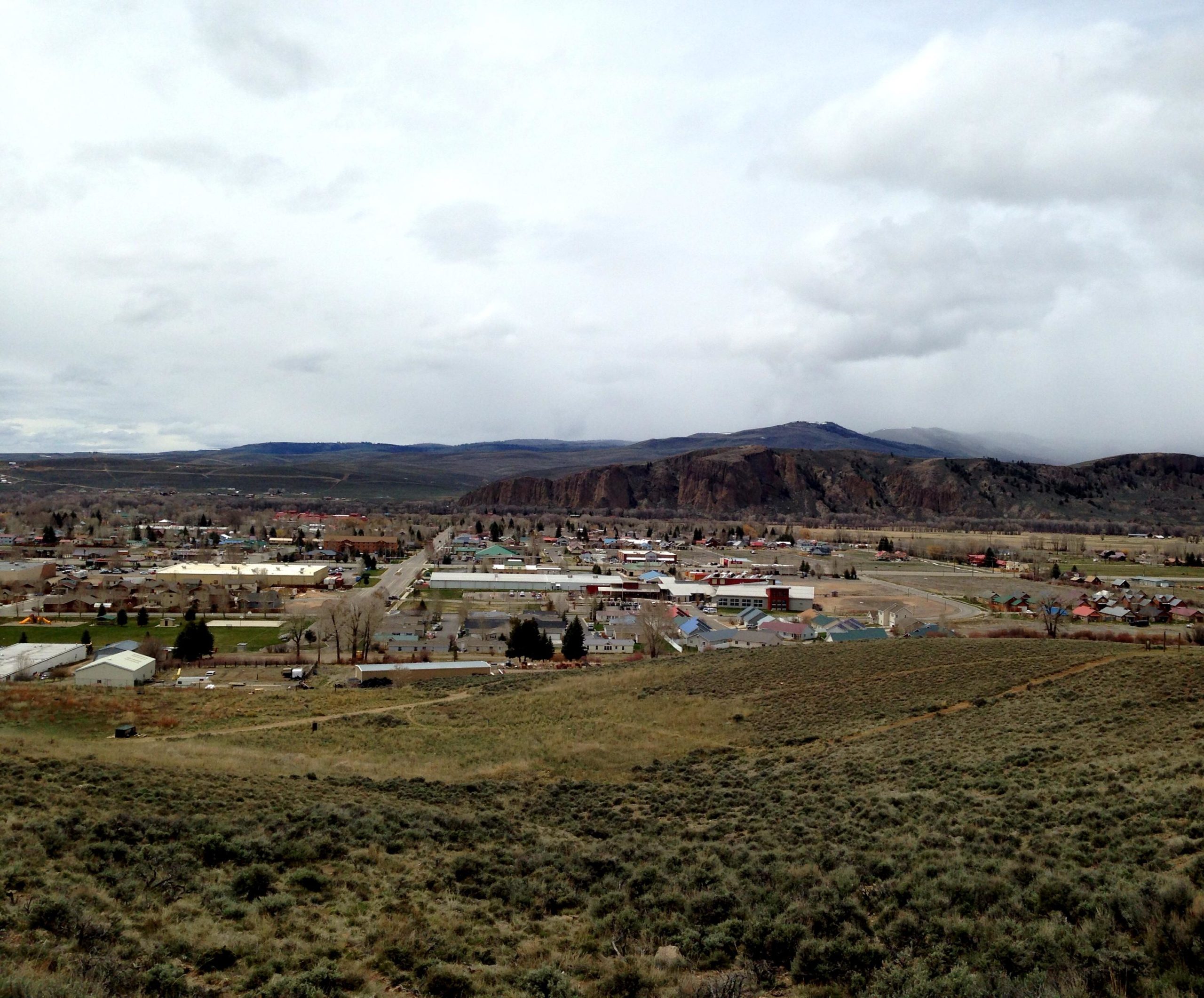 A panoramic view of a small town nestled in a valley, surrounded by rolling hills and mountains. The foreground features grassy terrain with sagebrush, while the town itself includes a mix of residential and commercial buildings. Overcast skies loom above, with distant mountains partially shrouded in fog. Signal Peak Trail System mountain bike trail.