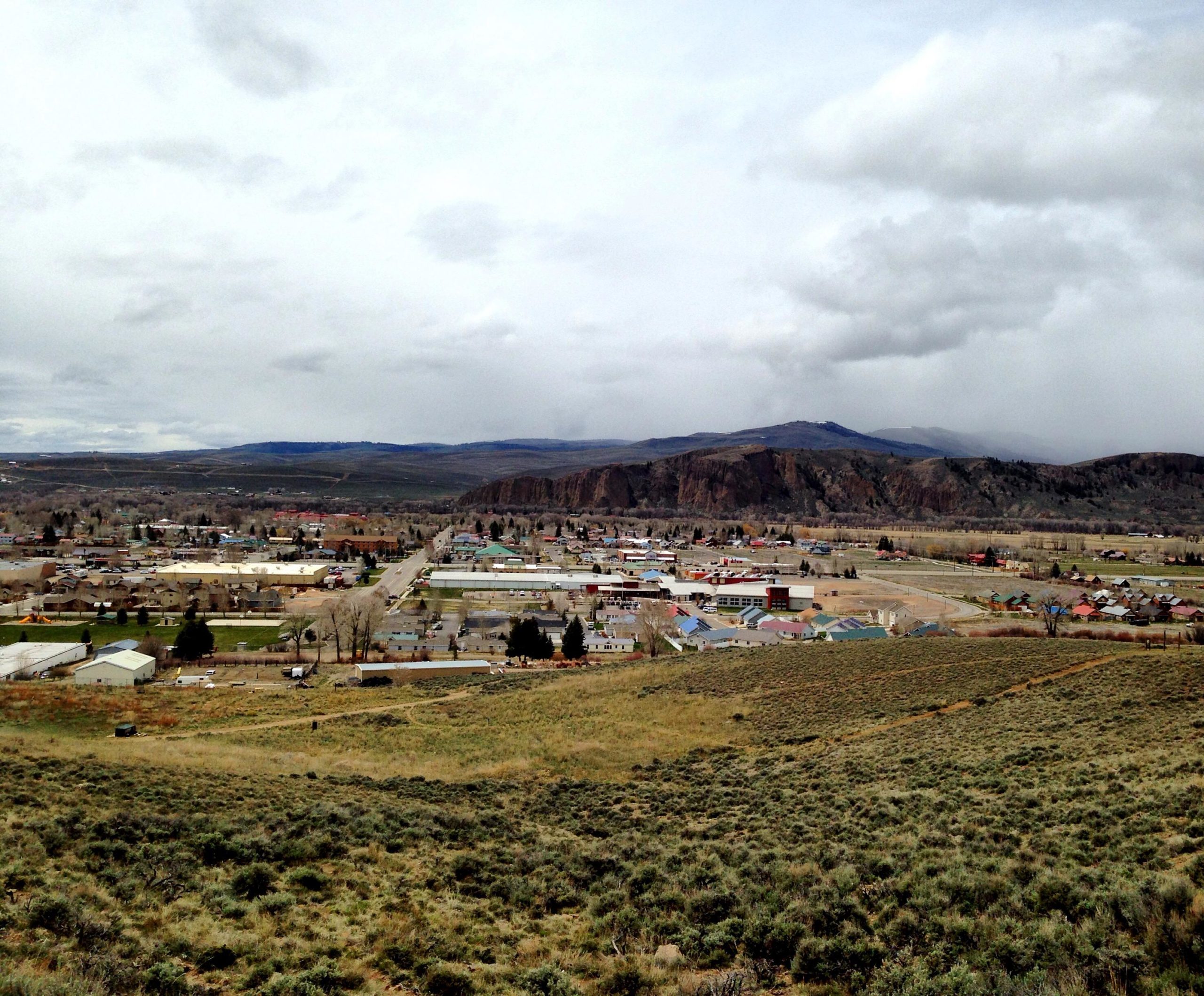 A panoramic view of a small town nestled in a valley, surrounded by rolling hills and mountains under a cloudy sky. The foreground features green shrubbery and open fields, while the town's buildings and streets stretch out below, showcasing various rooftops and structures amidst the landscape. Signal Peak Trail System mountain bike trail.