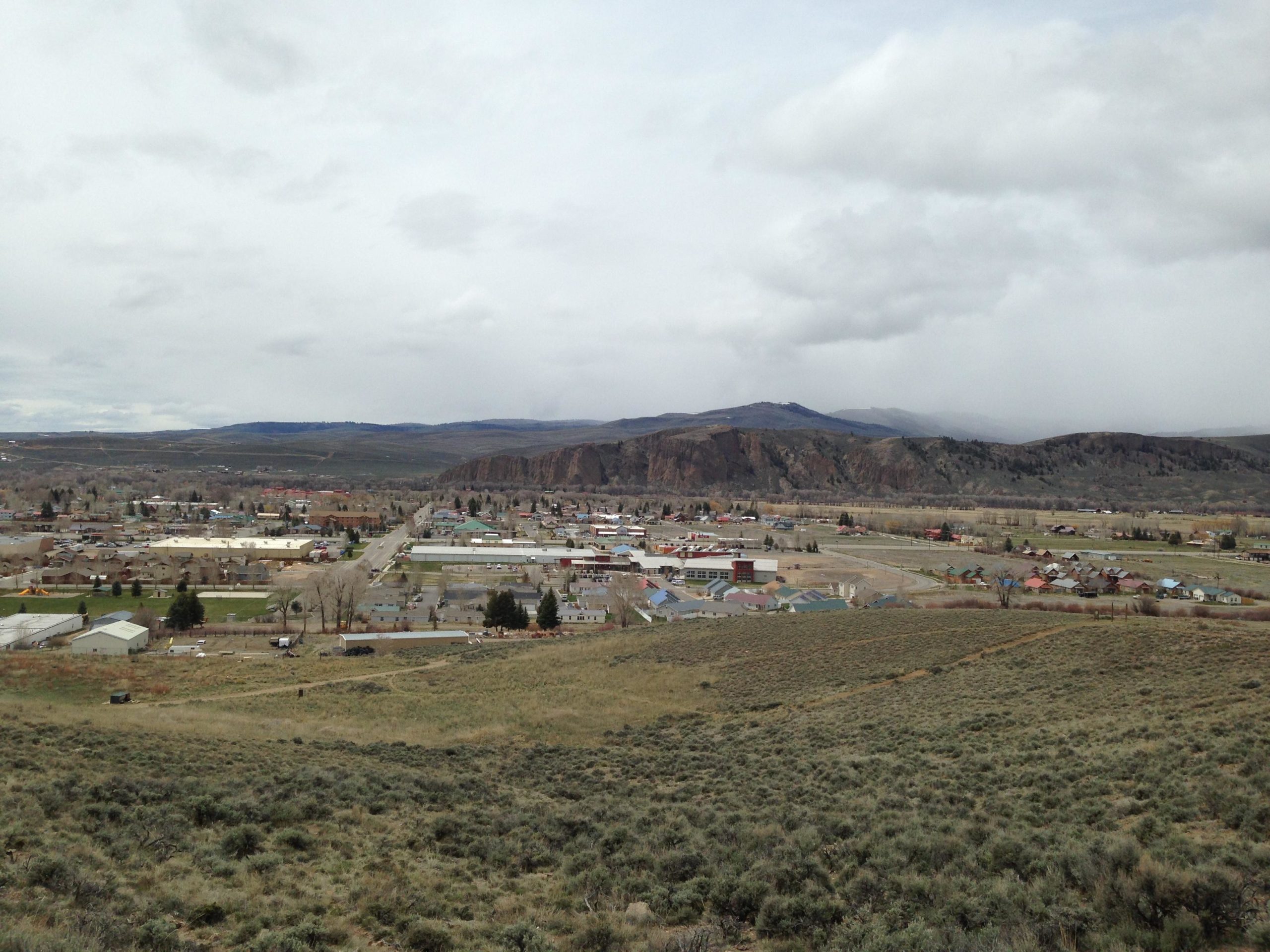 A panoramic view of a small town nestled in a valley, surrounded by rolling hills and distant mountains under a cloudy sky. The town features a mix of residential and commercial buildings, with tree lines and open fields visible. Signal Peak Trail System mountain bike trail.