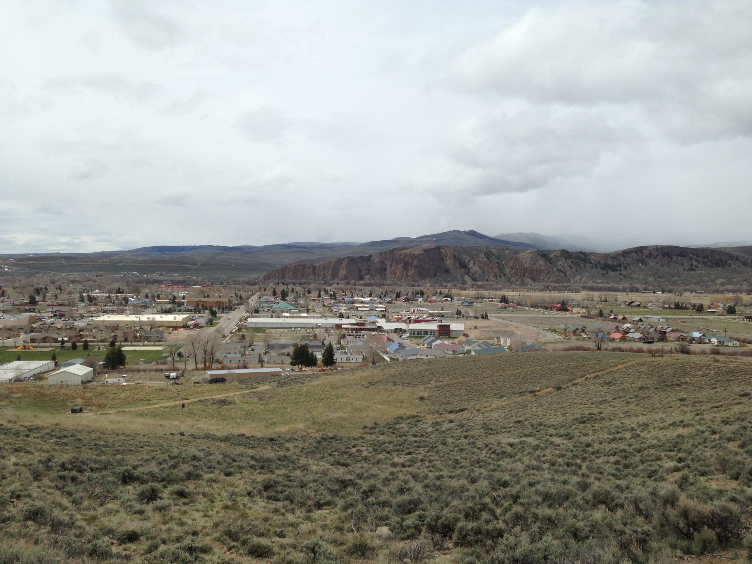 A panoramic view of a small town nestled in a valley, surrounded by hills and mountains under a cloudy sky. The foreground features grassy terrain and shrubs, while the town below displays a mix of residential and commercial buildings, roads, and patches of greenery. The distant mountains create a picturesque backdrop. Signal Peak Trail System mountain bike trail.