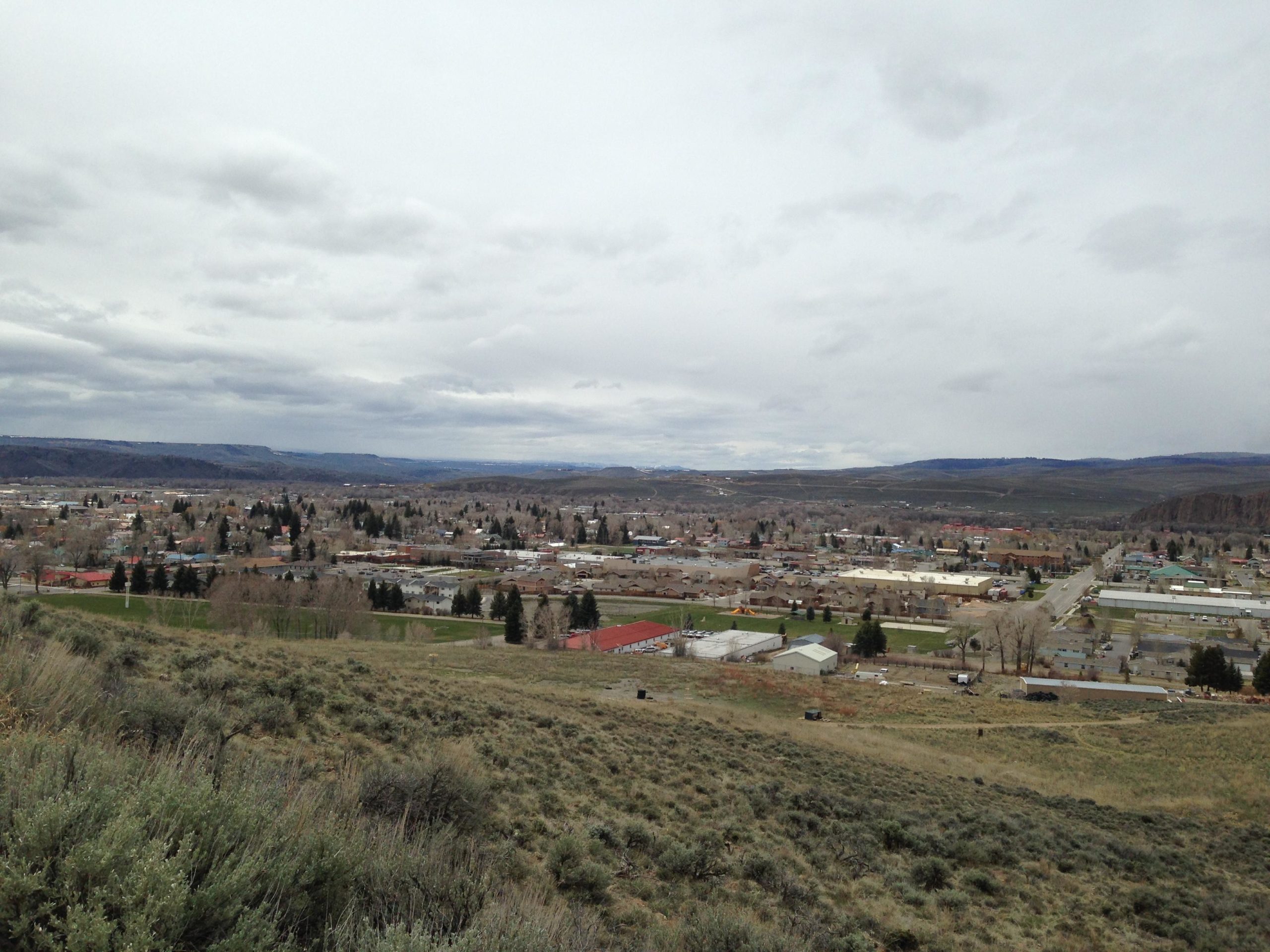 A panoramic view of a small town surrounded by rolling hills and mountains under a cloudy sky. The foreground features patches of grass and shrubs, while the town below includes a mix of residential houses and commercial buildings. The landscape is largely barren, with distant hills visible on the horizon. Signal Peak Trail System mountain bike trail.