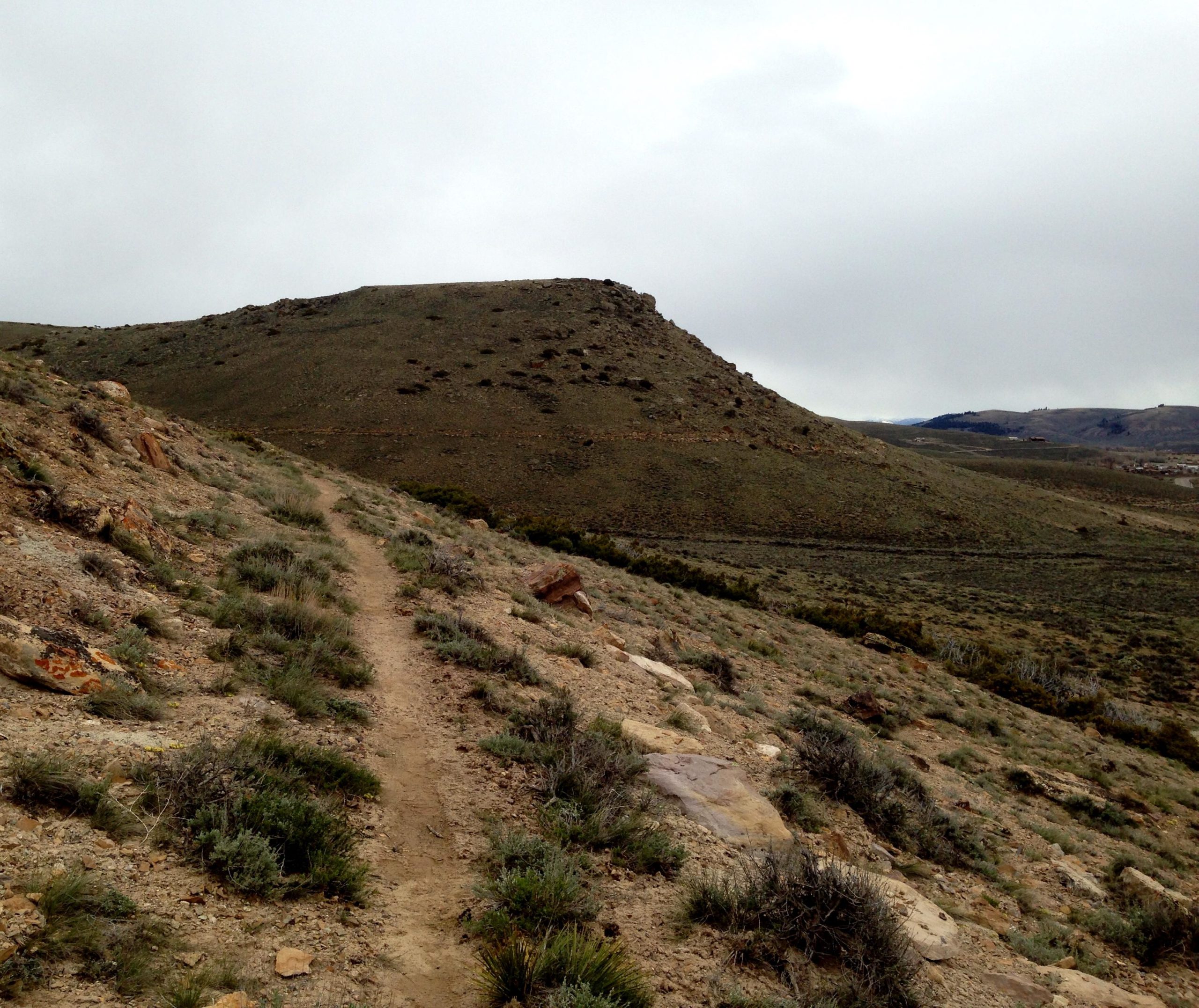 A dirt path winds along the side of a grassy hill, leading towards a higher elevation in the distance. The landscape features rocky terrain and sparse vegetation under a cloudy sky, creating a serene and isolated natural setting. Signal Peak Trail System mountain bike trail.