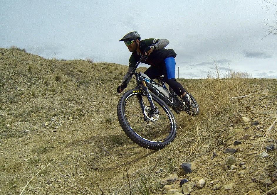A cyclist leans into a turn while riding a mountain bike on a dirt trail surrounded by sparse vegetation and a cloudy sky. The rider is wearing a helmet and sunglasses, showcasing dynamic movement as they navigate the terrain. Signal Peak Trail System mountain bike trail.