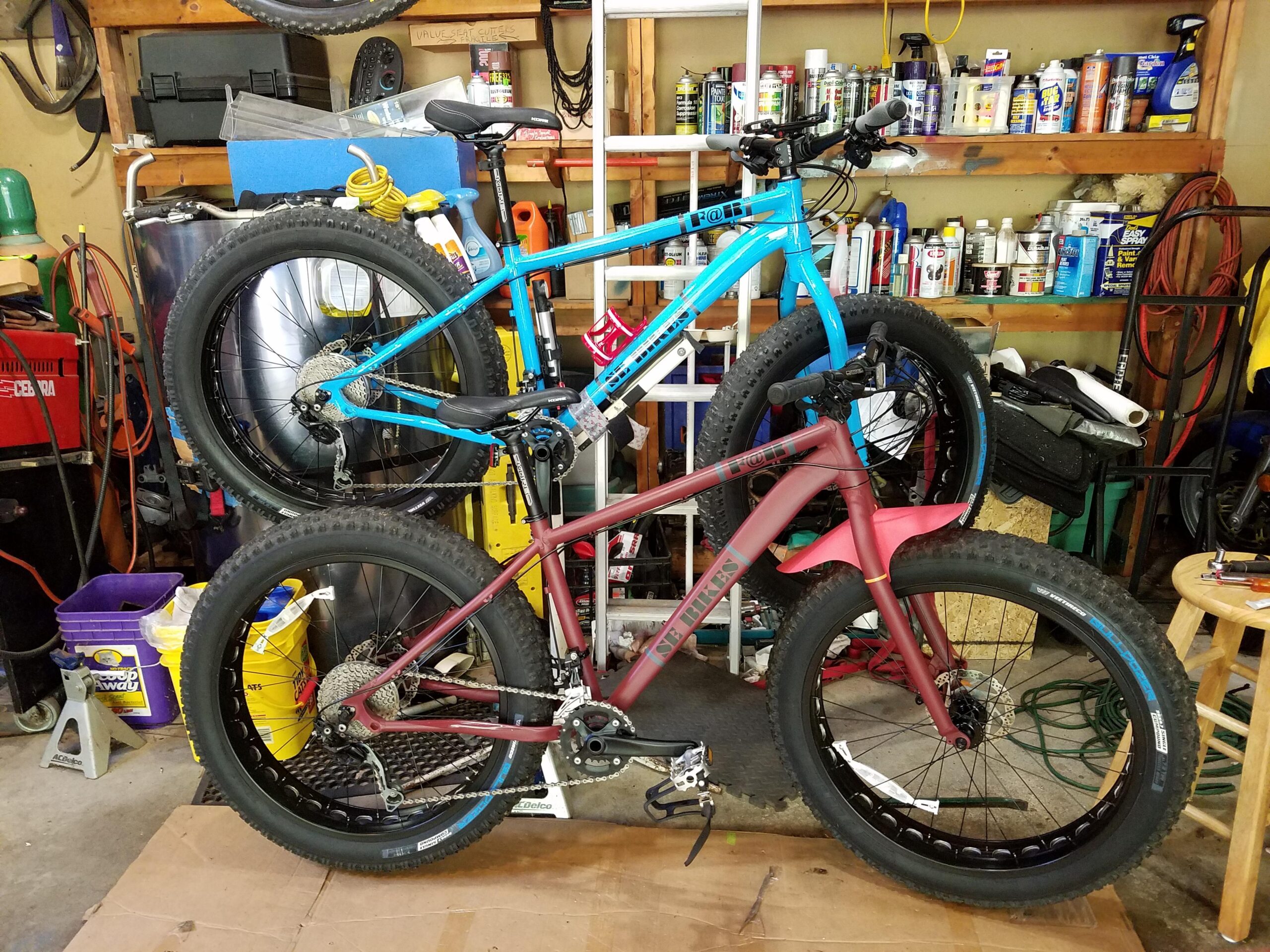 SE F@r: Two mountain bikes with oversized tires, one blue and one burgundy, are positioned vertically on a garage floor. The background features shelves filled with various tools and maintenance supplies, along with a ladder and other equipment. The bikes are surrounded by various items typically found in a garage setting.