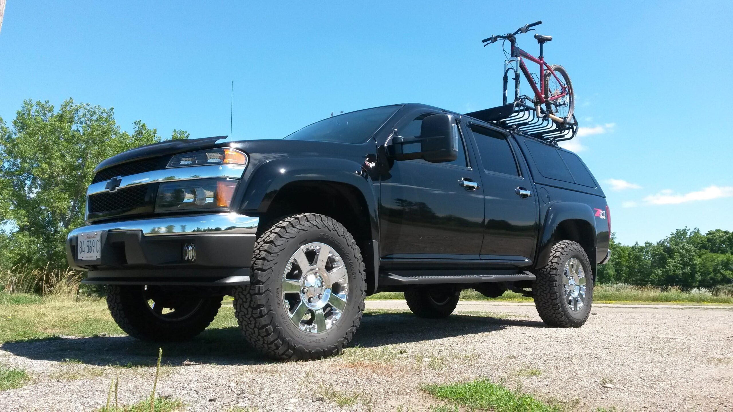 Specialized Hardrock Comp Disc: A black Chevrolet truck parked on gravel with a mountain bike secured on a roof rack. The truck features large chrome wheels and rugged tires, set against a backdrop of green trees and a blue sky.