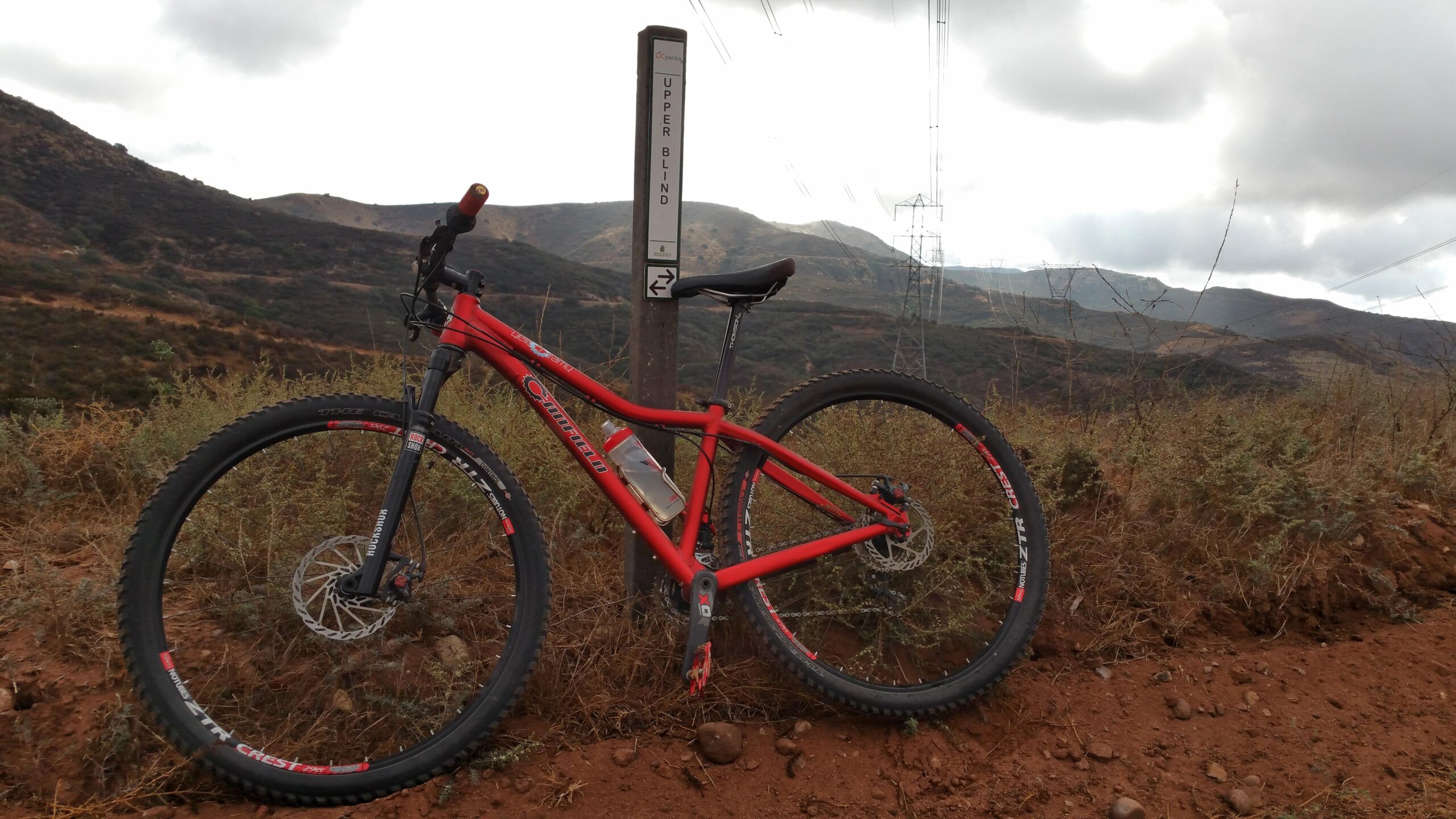 Canfield Brothers Yelli Screamy: A red mountain bike resting on a dirt trail next to a signpost labeled "Upper Blind," with mountains and cloudy skies in the background.