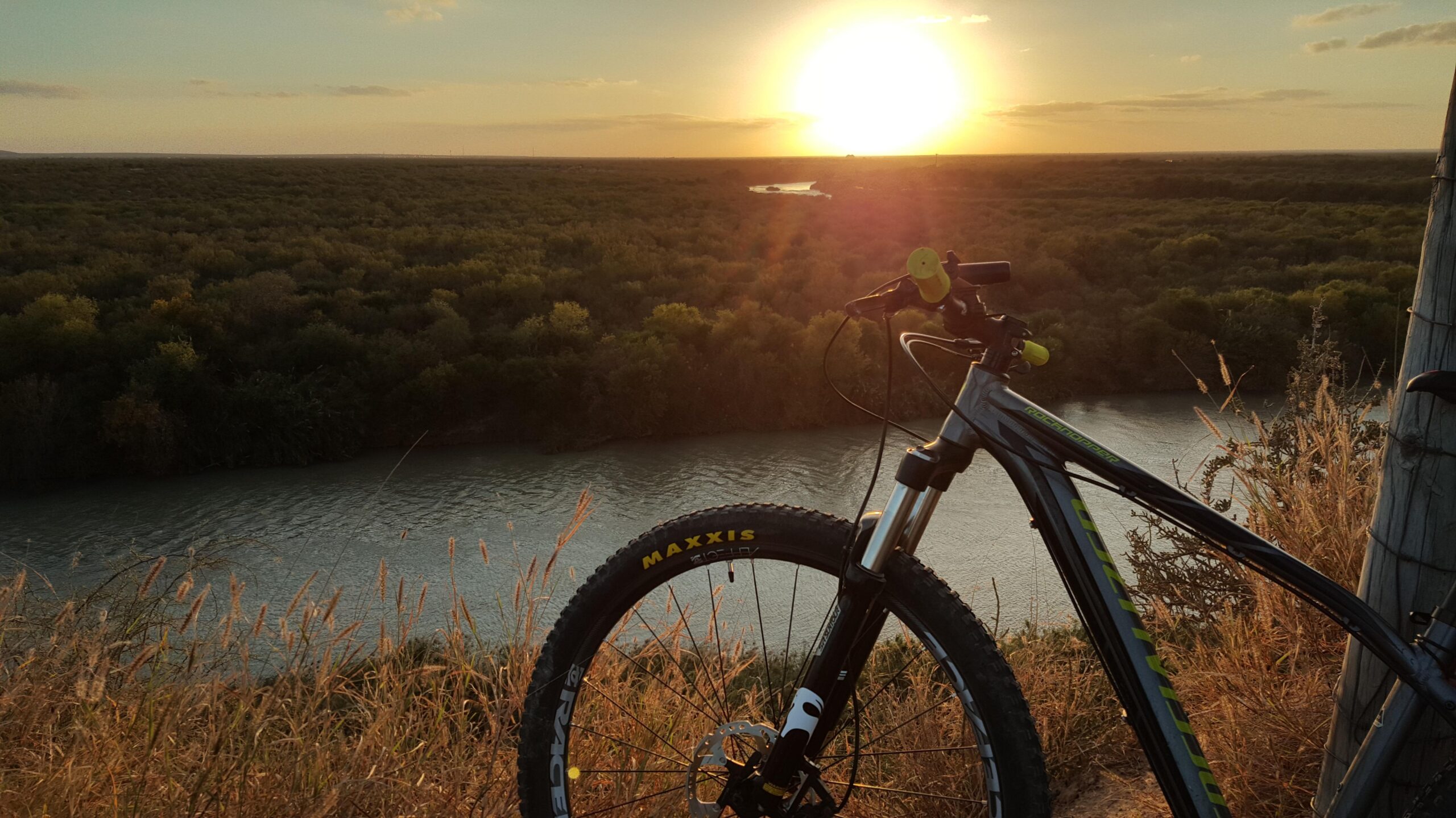 Specialized Rockhopper 29: A mountain bike resting on a grassy hillside, with a river flowing below and a vibrant sunset casting warm hues in the sky.