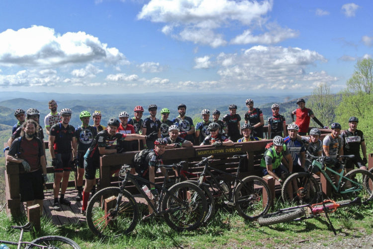 A group of mountain bikers poses for a photo at Mountaintown Overlook, with lush green mountains and a blue sky in the background. The riders wear cycling gear and helmets, and several bikes are leaned against a wooden railing.