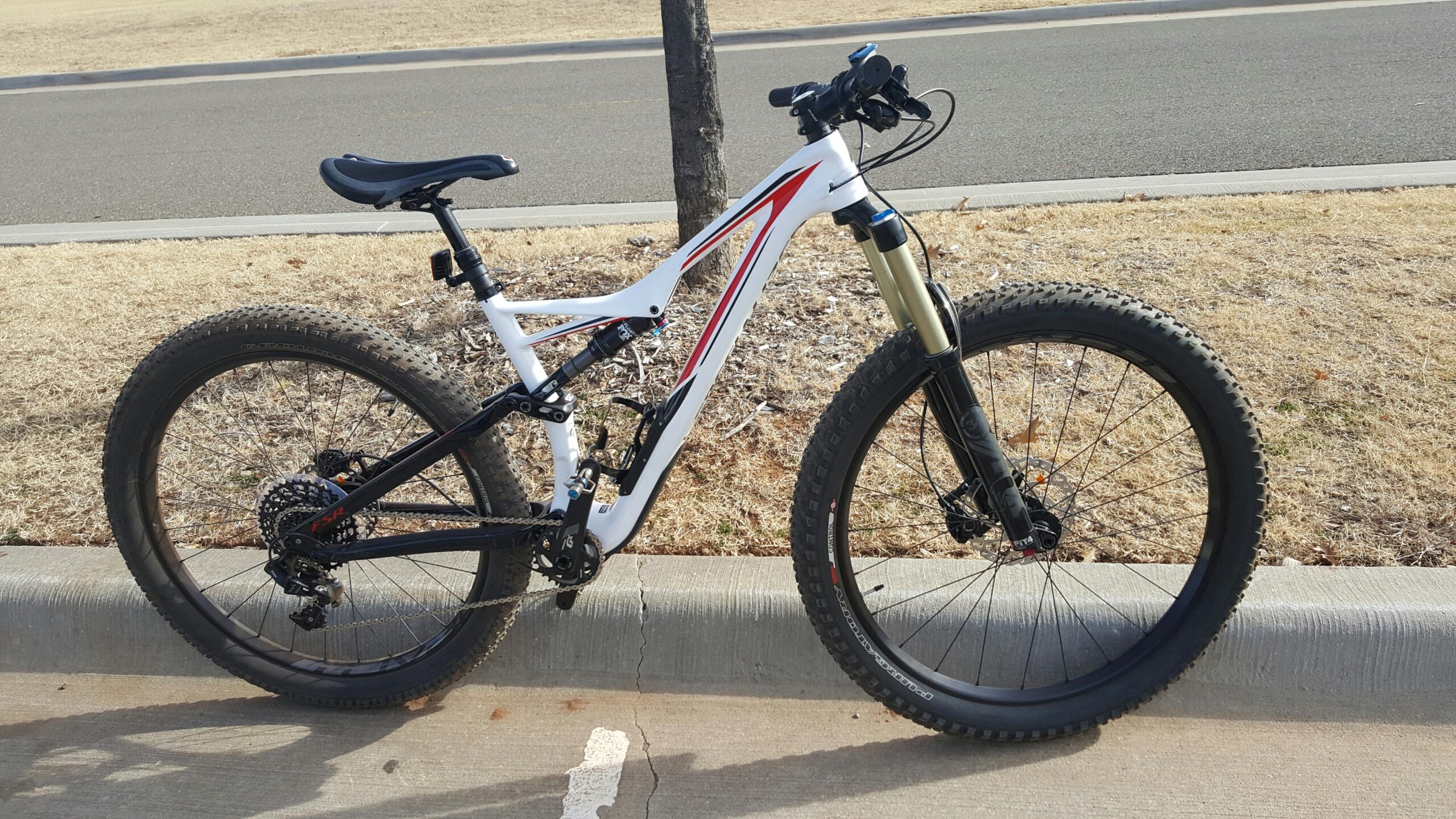 Specialized Stumpjumper FSR 6Fattie: A mountain bike with a white and black frame, featuring thick, knobby tires. The bike is parked on a concrete edge, with dry grass and a road in the background. The seat is raised, and the bike has a front suspension fork.