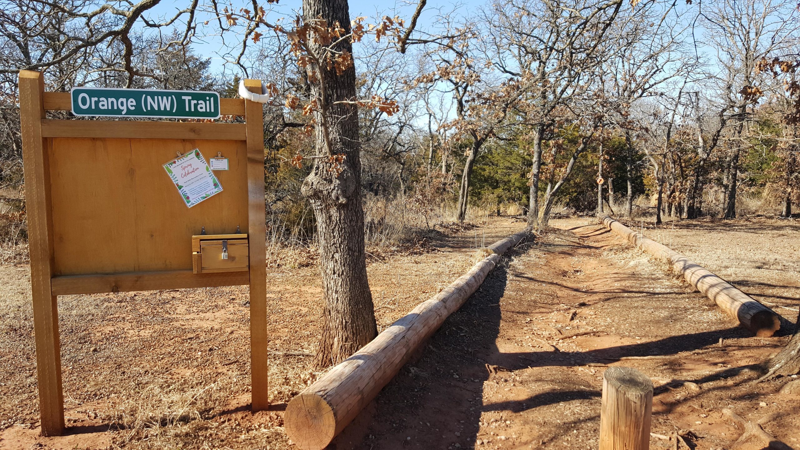 Sign for the Orange (NW) Trail at a natural park, featuring a wooden post with a bulletin board displaying announcements, surrounded by a dirt path lined with logs and trees. McMurtry Trail mountain bike trail.