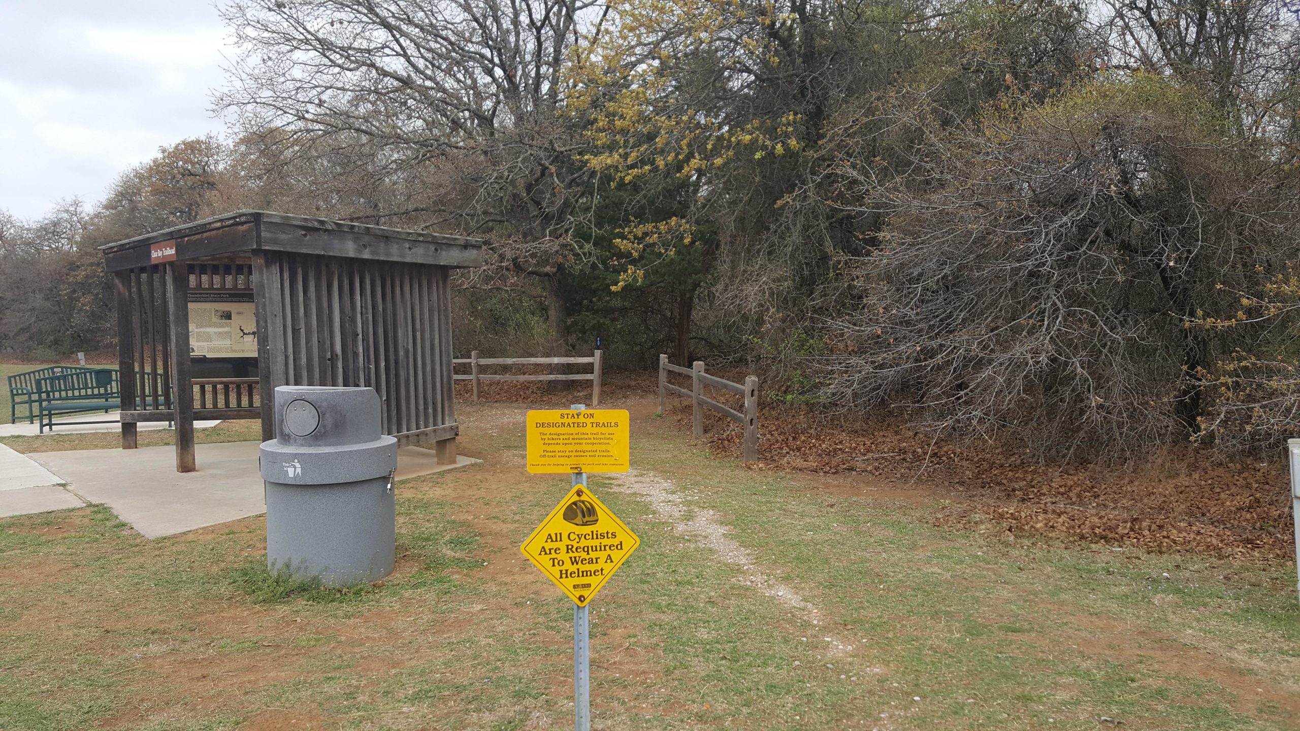A park area featuring a wooden shelter with informational displays, a green bench, a trash receptacle, and two signs: one advising visitors to stay on designated trails and another reminding cyclists to wear helmets. The surrounding landscape includes sparse trees and grass, with a cloudy sky above. Thunderbird Lake Clear Bay mountain bike trail.