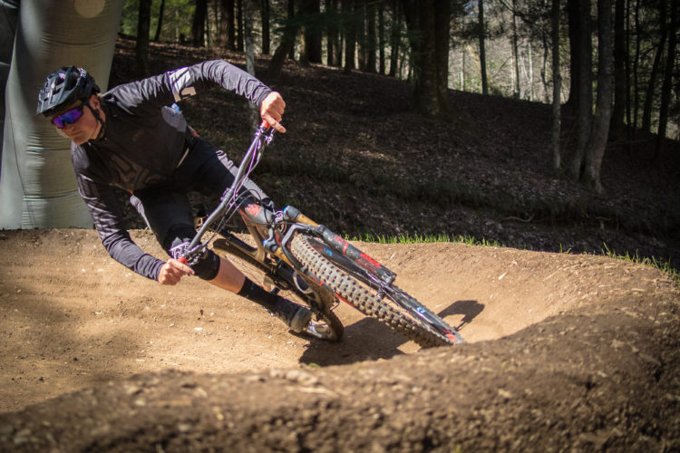 A mountain biker leans into a turn on a dirt track, wearing a helmet and sunglasses. The rider is focused and in motion, with trees and dappled sunlight in the background, showcasing a natural outdoor setting.