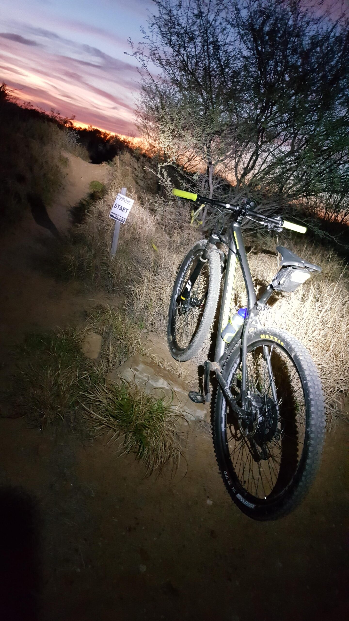Specialized Rockhopper 29: A mountain bike is parked next to a trail sign that reads "START" as the sun sets in the background, illuminating the sky with shades of purple and orange. The bike features a black frame with yellow accents and a water bottle attached, positioned on a dirt path lined with grass and small rocks, surrounded by sparse vegetation.
