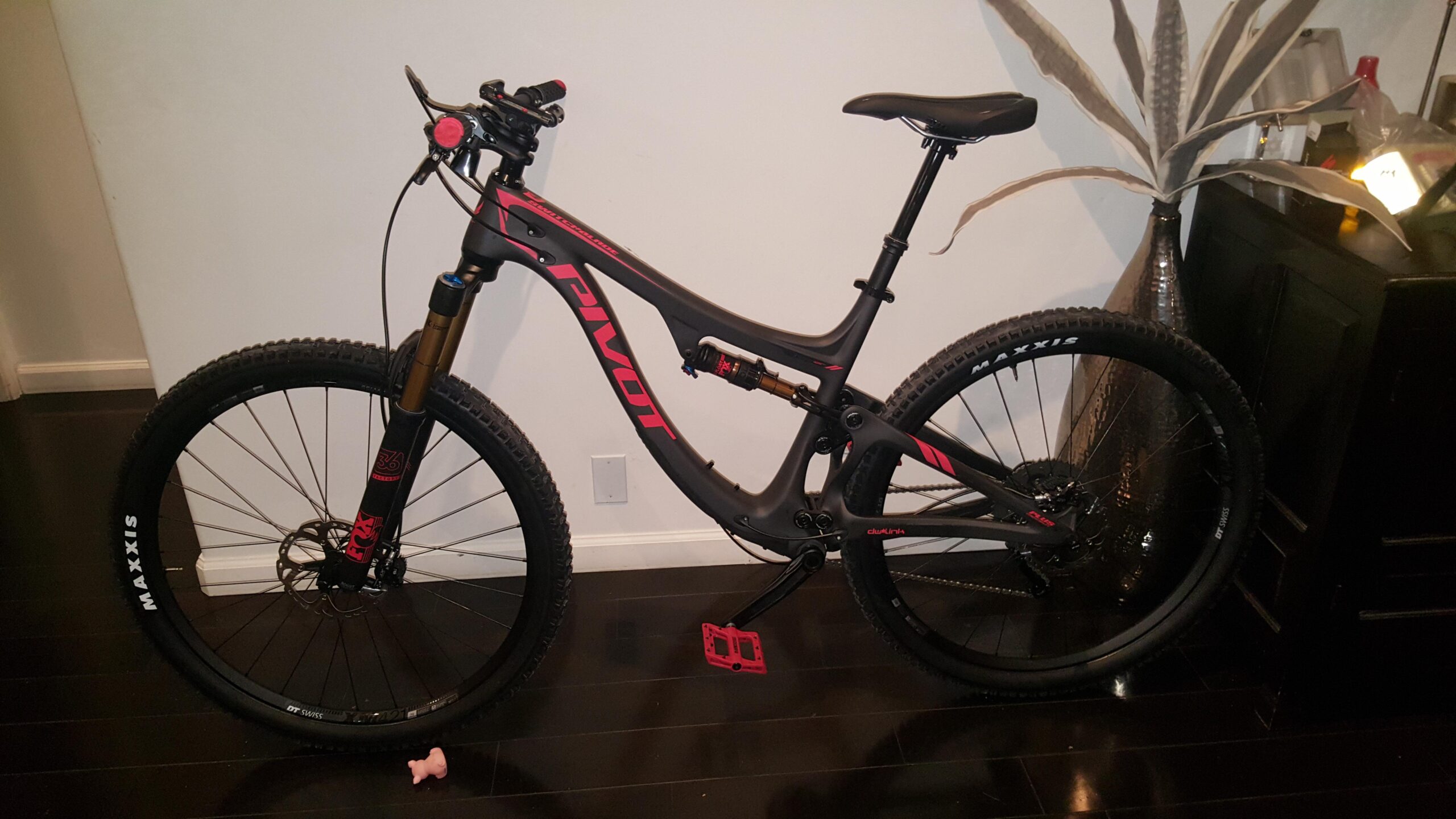 Pivot SwitchBlade: A black and red mountain bike leaning against a wall, featuring thick tires and a front suspension fork. The bike is positioned on a dark wooden floor, with a decorative plant and a piece of furniture in the background. A small pink object is visible in the foreground.