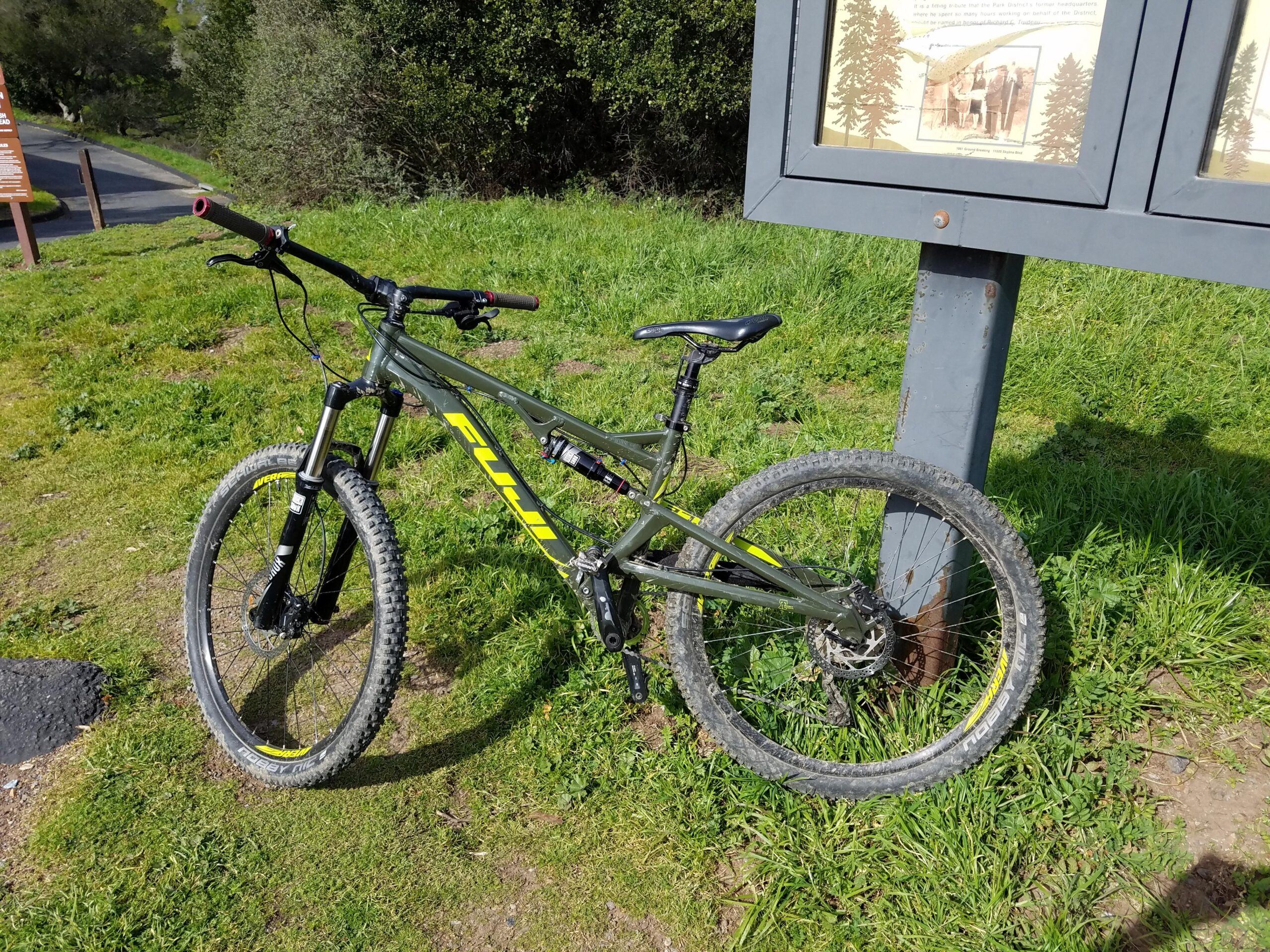 Fuji Fuji Reveal 1.1: A green mountain bicycle with yellow accents resting on the grass near a signpost in a natural outdoor setting. The bicycle features thick tires suitable for off-road terrain and is positioned upright next to a weathered informational sign. In the background, there are trees and a path leading into the distance.