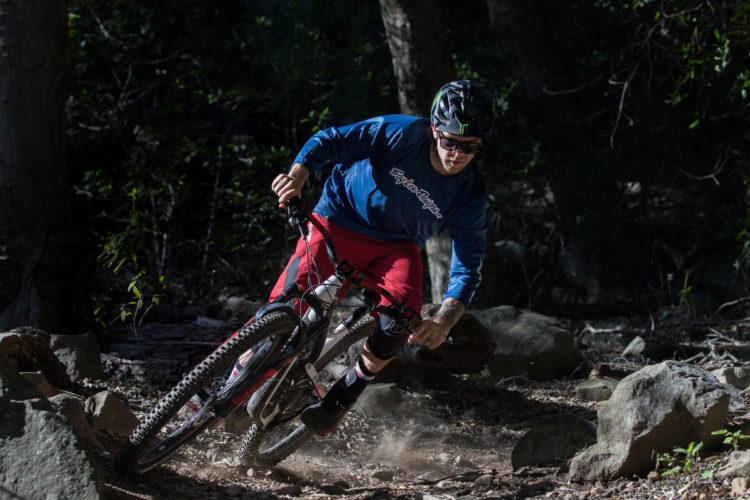 A mountain biker in a blue long-sleeve shirt and red shorts leans into a turn on a rocky trail, kicking up dust as he navigates through the rough terrain. Sunlight filters through the trees, highlighting the movement and energy of the ride.